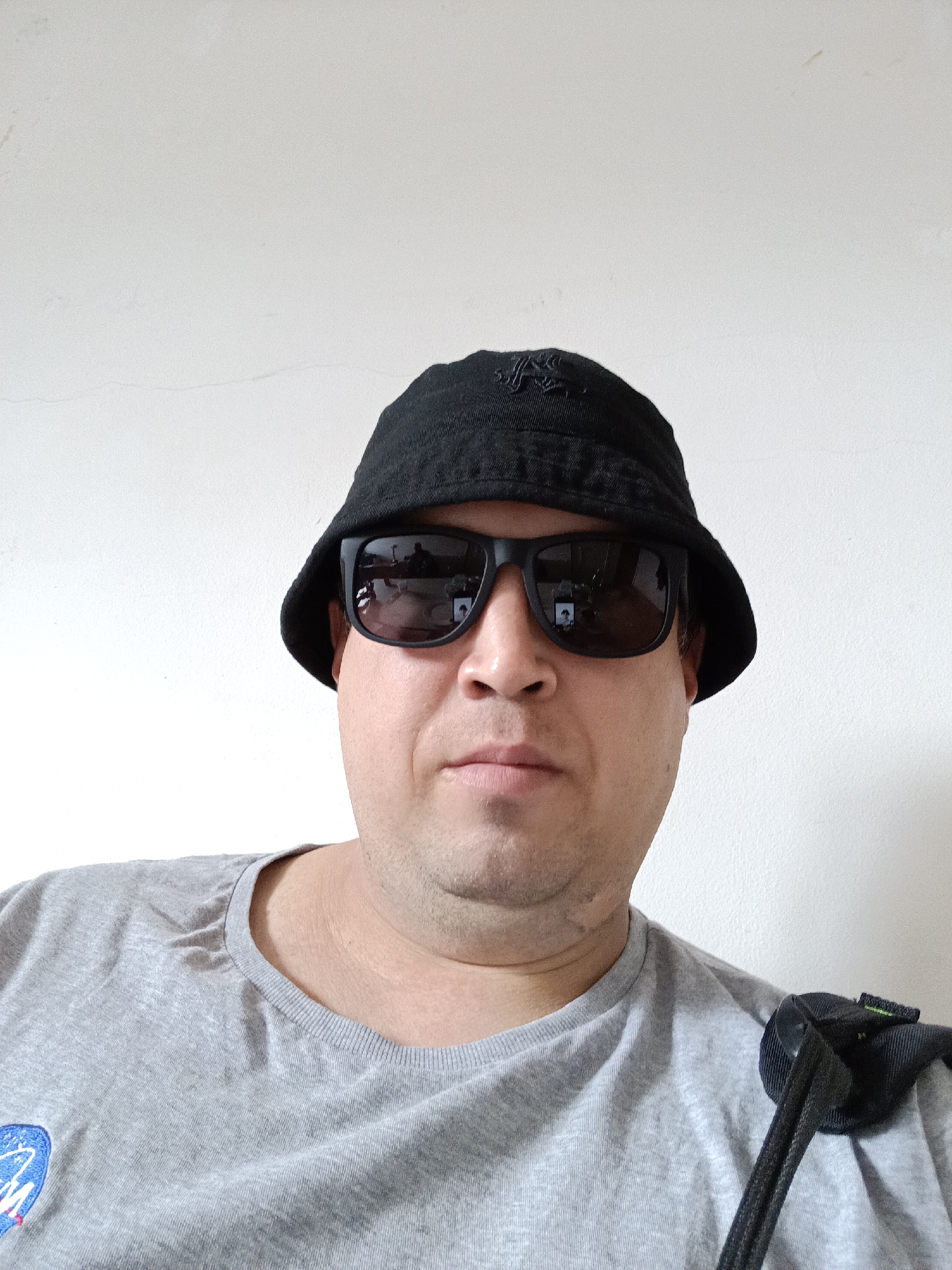 A head-and-shoulders portrait captures a man posing indoors against a plain white wall in Dalovy, Czechia. The man faces forward, looking directly at the camera with a neutral expression. He wears a black bucket hat and black sunglasses, which obscure his eyes. The sunglasses reflect an indoor environment, possibly showing the photographer and some room elements. He is dressed in a light grey, short-sleeved t-shirt. On the left side of his chest, a circular blue, white, and red logo is visible, which is clearly the NASA "meatball" emblem. A black strap, likely from a shoulder bag or backpack, rests over his right shoulder. The white background wall appears slightly textured with minor imperfections, including faint cracks and small stains. The lighting is even and soft, typical of an indoor setting.