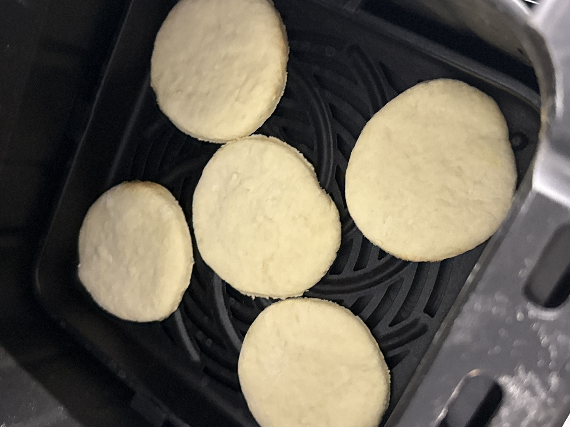 Five round, light-colored, uncooked pastries are arranged in a black air fryer basket. The pastries are spaced apart and do not touch each other. The air fryer basket has a ridged bottom. The image is a close-up shot, with the pastries filling most of the frame. The scene appears to be a kitchen. There is no information to suggest this scene is in Lyubertsy, Russia.