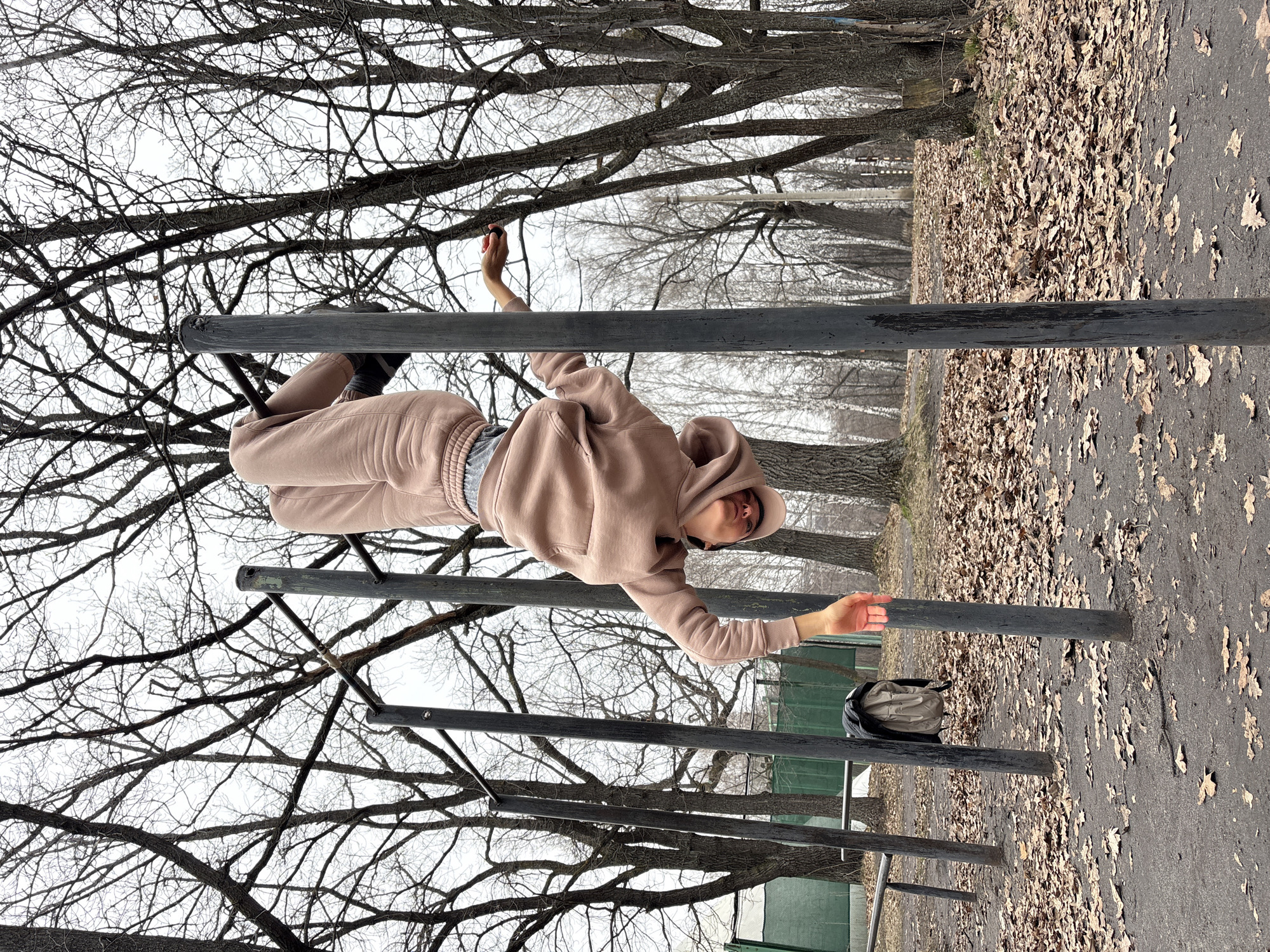 A woman wearing a beige hoodie and sweatpants is hanging upside down on parallel bars at an outdoor gym in Ulyanovsk, Russia. The ground is covered in fallen leaves, and bare trees surround the area. A backpack sits near one of the bars.