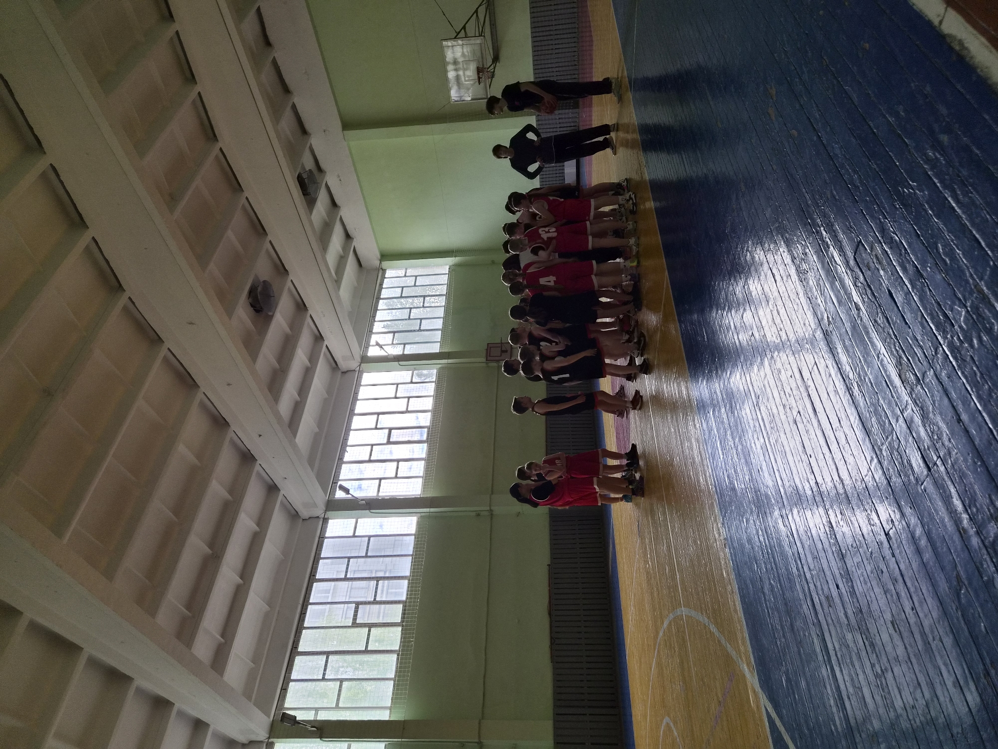 A basketball team stands in a huddle on a basketball court in a gymnasium in the city of Chaikovsky, Russia. The team wears red jerseys with black shorts, and they are being addressed by two coaches who are wearing black athletic wear. The gymnasium has a blue and yellow floor, green walls, and large windows that provide natural light. The ceiling has a grid-like design.