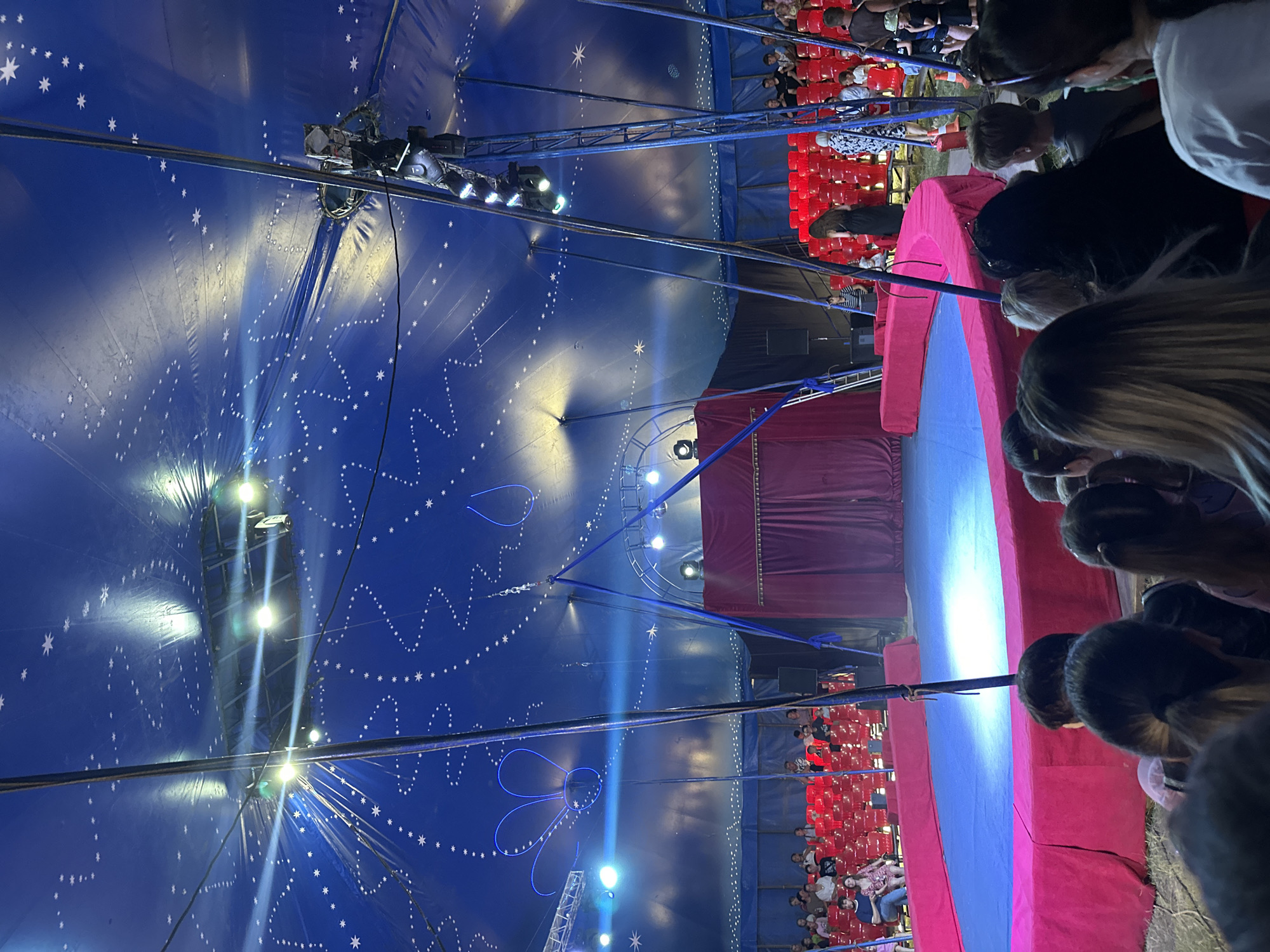 An indoor circus tent in Novoaltaysk, Russia, is shown from the perspective of the audience. The tent ceiling is a deep blue, adorned with numerous white star-like decorations and some larger, glowing blue outlines resembling flowers or celestial bodies. Multiple support poles and lighting rigs are visible throughout the tent.
In the center of the tent is a circular circus ring with a blue mat and a bright spotlight illuminating a section of it. The ring is bordered by a pink padded railing. Behind the ring, red velvet curtains hang on a stage-like structure.
Seating for the audience consists of rows of red chairs arranged around the perimeter of the tent. Some audience members, mostly children and adults, are visible sitting in these chairs and in the foreground. The people in the foreground are out of focus, suggesting the photo was taken from within the seated audience area.
Several individuals are standing near the ring and on elevated platforms, possibly performers or circus staff. The overall atmosphere appears to be that of an ongoing circus performance or a pre-show setup. The lighting is dim and theatrical, with spotlights creating bright beams.