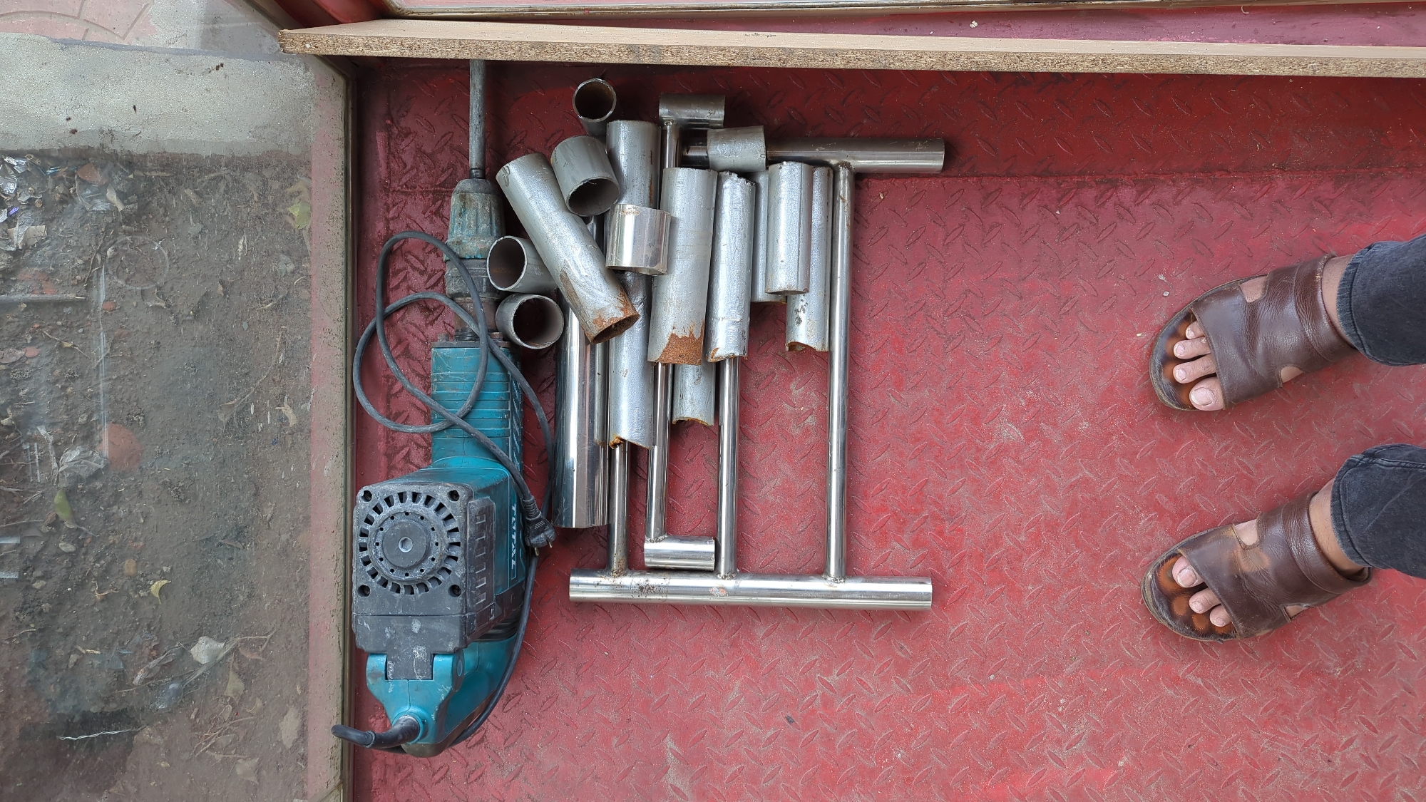 The scene is an overhead shot of a metal flatbed in Dhaka, Bangladesh, with a person's feet visible in the lower right corner. The bed contains an electric jackhammer, several sections of silver metal pipe, and one assembled "T" shaped metal fitting. The surface of the metal flatbed is red with a diamond plate texture. The electric jackhammer is turquoise and black. The pipe sections are of varying lengths and show some corrosion. The person is wearing brown leather sandals and black jeans. The background is a mix of concrete and wood framing.