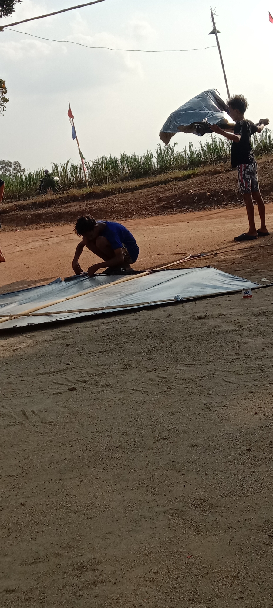 Two young males are in an outdoor environment, likely a field or unpaved area. One boy, wearing a dark t-shirt and patterned shorts, stands to the right, holding a large, light-colored fabric that appears to be part of a kite, aloft. He is looking towards the fabric. The second boy, wearing a blue t-shirt and dark shorts, is crouched down on the left, assembling a large, flat object that also seems to be a kite. This object is laid out on the ground, with bamboo poles forming its frame. The background shows a line of tall green plants, possibly crops, and a dirt road. In the distance, a motorcycle is visible. The sky is bright, indicating daytime. The overall scene suggests preparations for flying a kite. The location is identified as Utama Jaya Mataram, Indonesia.
