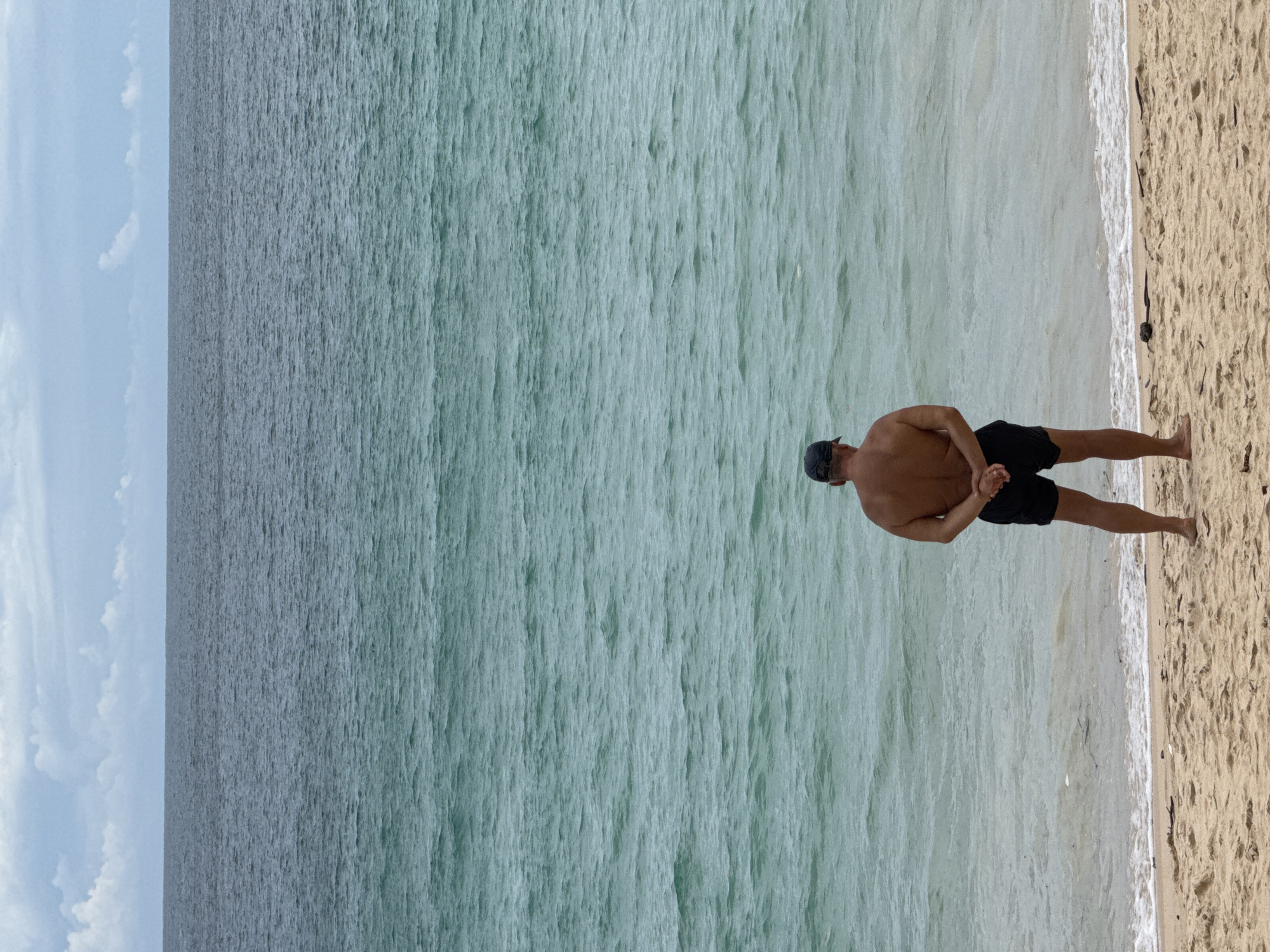 A man stands on a sandy beach in Rawai, Thailand, facing the turquoise ocean. He wears black swim shorts and a dark baseball cap. His hands are clasped behind his back. The water is calm with gentle waves reaching the shore. In the background, the sea stretches to the horizon, meeting a cloudy sky. The sand is light brown with scattered debris.