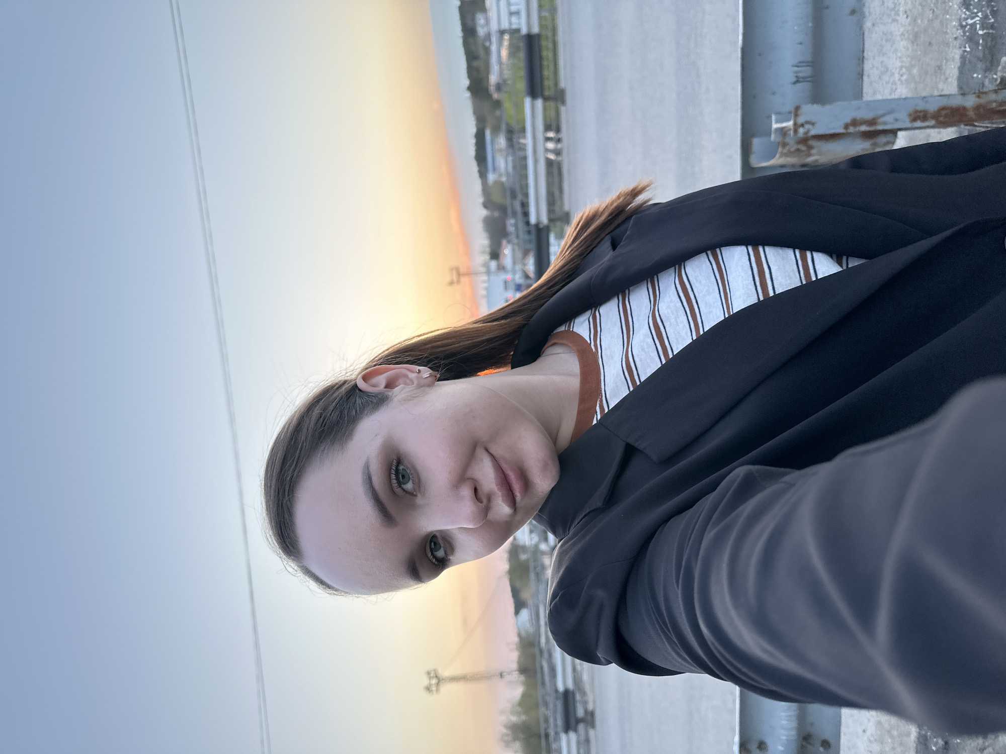 A selfie shows a woman with light skin, hazel eyes, and brown hair pulled back, wearing a striped shirt and black blazer. The background features a sunset gradient in the sky, transitioning from blue to orange. Faint overhead power lines are visible in the sky. A gray concrete structure is in the foreground. The background cityscape includes industrial elements and trees, suggesting a location within the city of Chaikovsky, Russia.