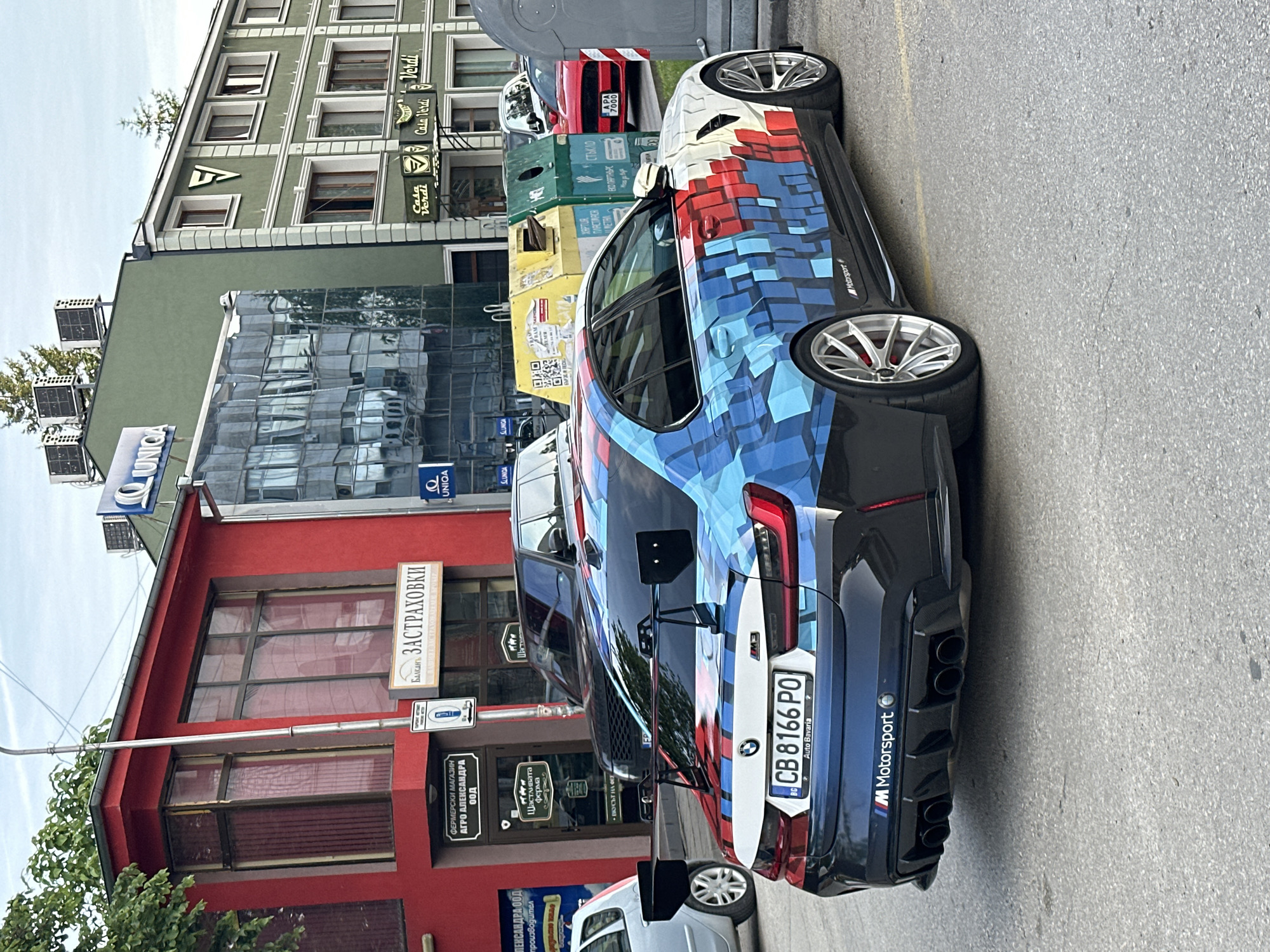The image shows a custom-wrapped BMW M3 parked on a street, likely in Veliko Tarnovo, Bulgaria, as evidenced by the Cyrillic script on nearby signs and the Bulgarian license plate "CB 8166 PO". The car's wrap features a pixelated design in red, white, and blue. Behind the car are several buildings: a red building with signs for "АГРО АЛЕКСАНДРА" and "ЗАСТРАХОВКИ", a glass-fronted building with "UNIQA" signage, and a green building with "Casa Verdi" branding. Recycling bins are visible next to the car. Other cars are parked nearby.
