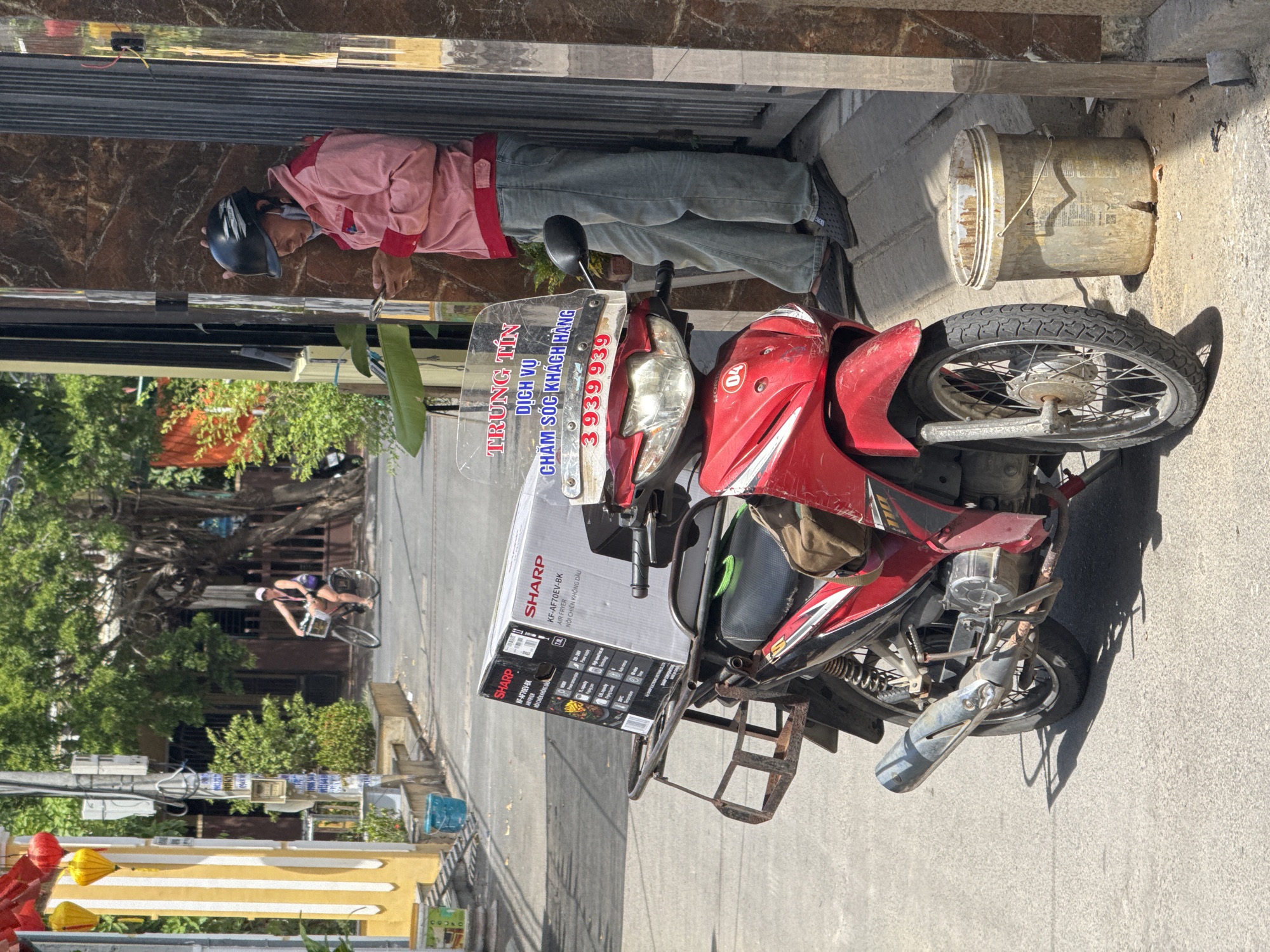 This is a medium shot, eye-level photograph of a man standing next to a red motorcycle on a street in Hoi An, Vietnam. The man, wearing a pink jacket, blue jeans, and a black helmet, is looking down at his phone. The motorcycle, parked on the right side of the street, has a large box from SHARP on its rear rack. The box appears to contain an air fryer. The street is paved with asphalt and lined with buildings on both sides. The left side of the street features a yellow building with white vertical stripes and hanging lanterns. Further down the street, a woman on a bicycle is visible. The overall environment suggests a typical street scene in a Vietnamese city, with motorcycles being a common mode of transportation and businesses operating from street-level shops. The light indicates it is daytime, with shadows cast by the buildings and objects.