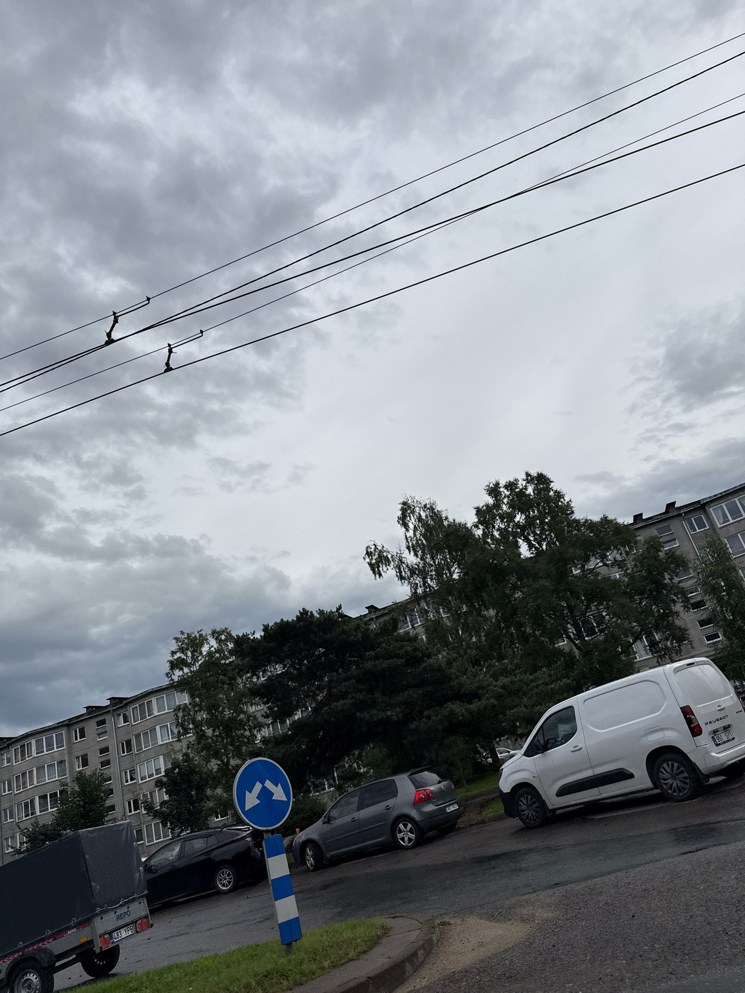 The image shows a cloudy sky over a street in Tallinn, Estonia, with buildings and cars visible. A long, multi-story apartment building runs along the left side of the image, with many windows. In front of the building, trees are growing, some tall and leafy, others bare. Several cars are parked on the street, including a white van on the right and a dark hatchback in the middle. A blue and white traffic sign with two arrows indicating left and right turns is positioned on the right side of the street. A trailer is parked on the left side of the street. Overhead power lines are visible against the sky.