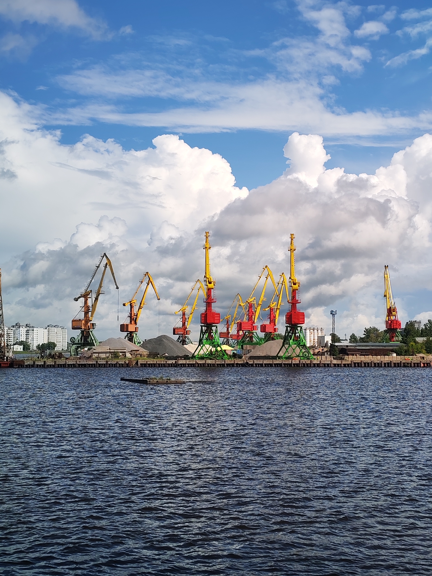 A wide-angle, eye-level outdoor photograph depicts a bustling port scene under a partly cloudy sky. In the foreground, dark blue, rippling water dominates the lower half of the frame. A long, dark, rectangular object floats on the water's surface towards the center. The mid-ground features a concrete pier lined with large, industrial-style cranes. These cranes are a mix of red, yellow, and green, with some having tall, red counterweights. Piles of gravel and sand are visible behind the cranes. In the background, a row of white, multi-story buildings stands out against the sky, suggesting an urban setting beyond the port. Trees line the far right side of the scene, interspersed with industrial structures. The sky is a vibrant blue with numerous white, fluffy clouds. The overall impression is of an active, industrial waterfront.