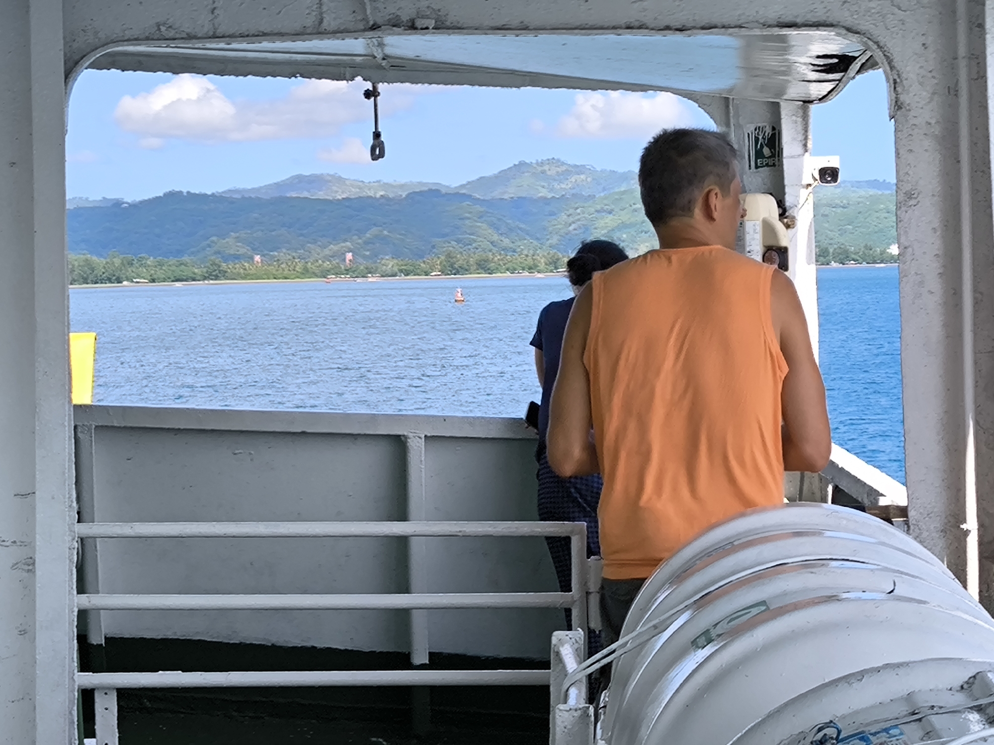The image captures a tranquil daytime scene from the deck of a boat or ferry, looking out over calm waters towards a distant mountainous coastline. In the foreground, the grey metal framework of the vessel creates a portal-like view, with white railings visible at the bottom left.
Two individuals are seen from behind, gazing out at the scenery. A man wearing a bright orange sleeveless shirt is prominent on the right, with another person partially visible to his left, both observing the view. On the far right, a large, white, cylindrical container, likely housing a life raft or similar safety gear, is secured to the boat's structure.
The expanse of water is a deep blue, reflecting the clear sky. A red and white buoy floats in the mid-distance. Beyond the water lies a lush green shoreline, densely forested with tropical trees, and subtle hints of structures along the coast. Majestic, rolling mountains, covered in thick vegetation, form the backdrop under a vibrant blue sky dotted with scattered white clouds.
On the boat's interior frame, to the right, a security camera is mounted, and below it, a white sign bears the partially visible text "EPIR". The bright, even lighting indicates it is daytime with clear weather conditions. The tropical landscape, clear waters, and mountainous islands align with the provided context of Lembar Selatan, Indonesia, suggesting a ferry journey through a scenic archipelago.