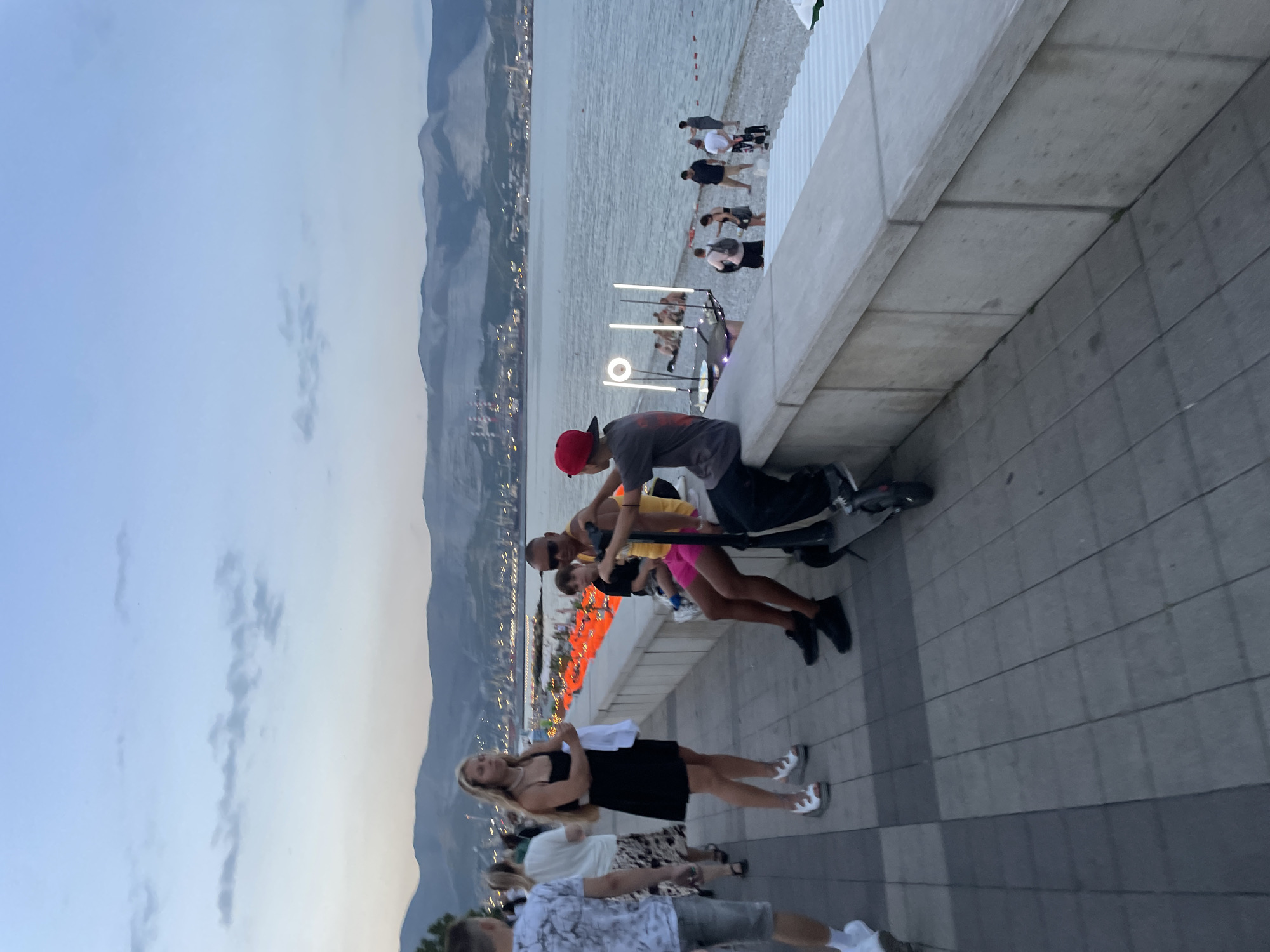 The image captures a scene along the embankment of Novorossiysk, Russia, near the city's port area, during the twilight hours. In the foreground, a paved walkway runs alongside a low concrete wall overlooking the sea. Several individuals are present. To the left, a woman in a black dress and white platform sandals stands with a white shirt in her hands. Further down the walkway, a man and a woman are seated on the wall, accompanied by a boy. Another young person in a red cap is seated on an electric scooter, facing away from the camera.
The sea is calm, and across the water, the lights of the port and the distant mountains are visible, shrouded in the dim evening light. A series of orange buoys or floating markers are present in the water near the shore. Other people are scattered along the beach and walkway in the background. The sky is a pale blue with some wispy clouds, indicating the end of the day. The overall atmosphere suggests a relaxed evening gathering by the sea.