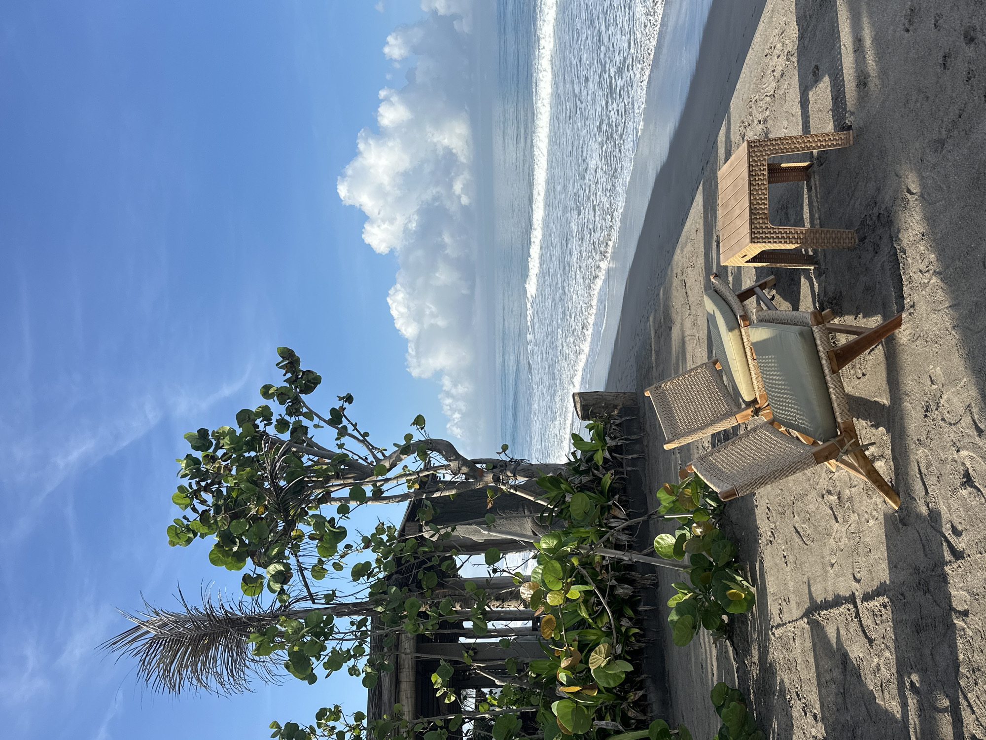 The image depicts a beach scene in North Kuta, Indonesia. A weathered beach chair and small side table sit on dark sand near the shoreline. The ocean is visible with gentle waves breaking. Behind the chair and table, there is a decaying wooden structure partially obscured by foliage. A tree with large, round leaves is prominent in the foreground. The sky is blue with some puffy white clouds. There are no people visible.