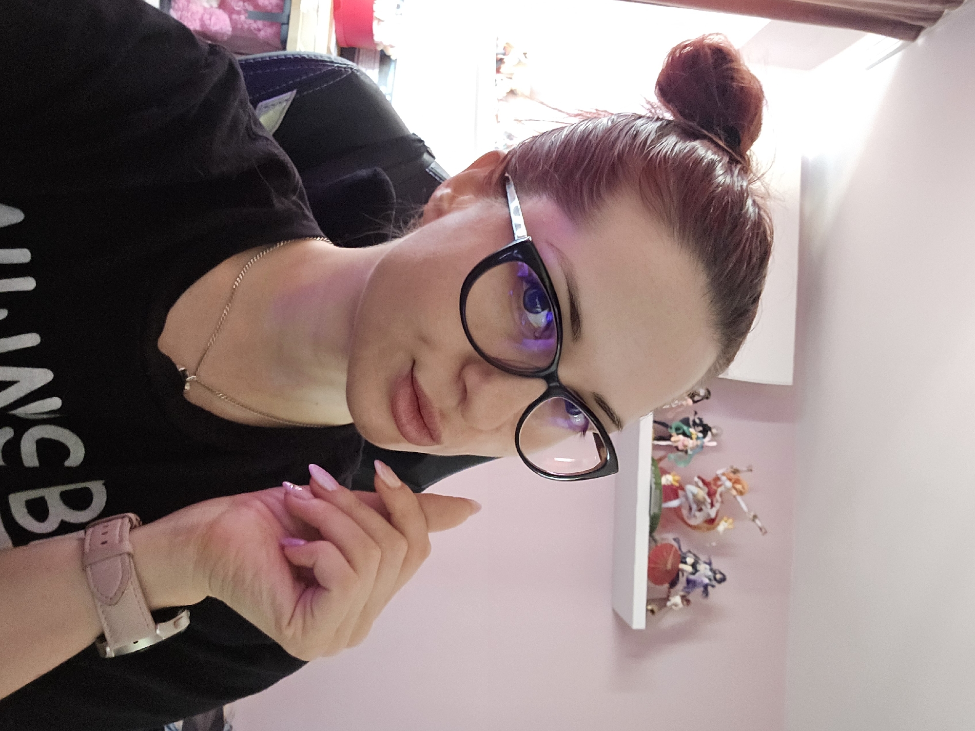 A woman with auburn hair in a bun, wearing black-rimmed cat-eye glasses, a black t-shirt, and a silver necklace, is captured in a close-up selfie. Her left hand is raised to her face, with light pink painted nails. A light pink watch is visible on her left wrist. Behind her, a white shelf holds several anime figurines against a pink wall. The background also shows a window with curtains and a glimpse of a pink storage bin and a black chair. The lighting suggests an indoor setting. The image does not provide any information about the city of Burgas, Bulgaria.