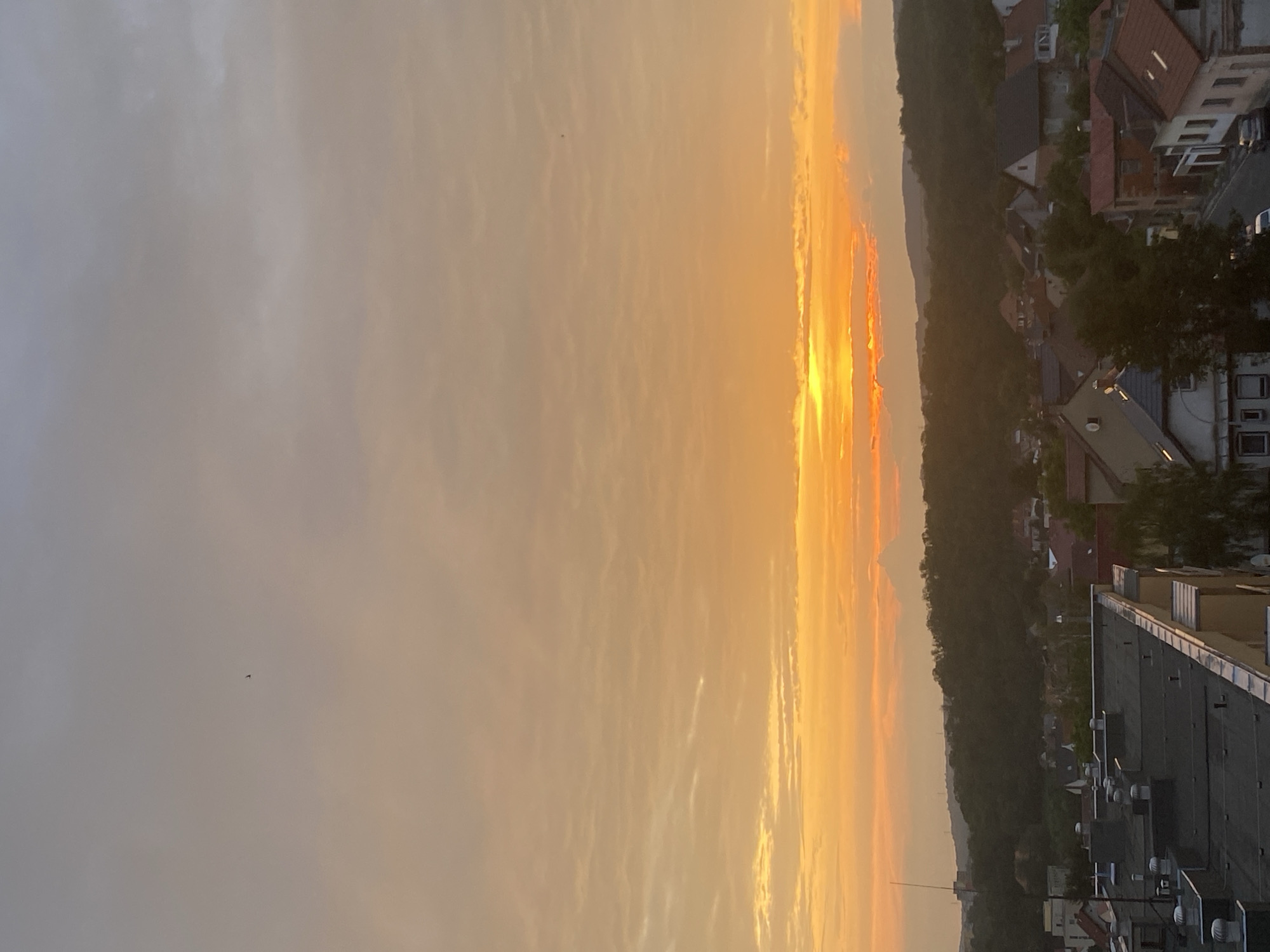 A high-angle, wide shot shows a silhouetted view of the town of Bílina, Czechia, during a vibrant sunset. The sky is filled with a warm gradient of orange and yellow hues, with scattered clouds creating texture. The sun is visible near the horizon, partially obscured by clouds, casting a golden glow over the landscape.
In the foreground, the flat roofs of several multi-story buildings are visible, characterized by dark grey surfaces and numerous white vents or pipes. A faint antenna or pole extends upwards from one of the buildings on the left. Beyond the buildings, a dense line of dark green trees forms a natural backdrop, suggesting a hilly or mountainous terrain. Nestled within this greenery and extending towards the foreground are numerous houses with red and brown tiled roofs, typical of European architecture. Some houses appear to be under construction or renovation, with exposed brickwork and incomplete structures. A street is visible winding through the houses, with a few parked cars. The overall impression is a serene, rural or suburban landscape at dusk. In the upper left quadrant of the sky, a small, dark silhouette of a bird is visible in flight.