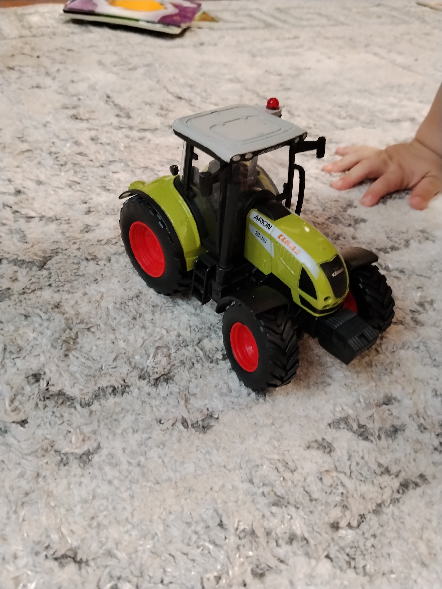 An overhead, slightly angled medium shot shows a green and grey toy tractor on a cream-colored, textured carpet. The tractor has large black wheels with red hubs. The text "ARION 540" and "CLAAS 12" are visible on the side of the tractor's hood. A small red beacon light is on top of the tractor's cab. To the right of the tractor, a child's hand is reaching towards it. In the background, slightly out of focus, is a colorful object, possibly a toy or book. The environment appears to be an indoor space, likely a home. There is no identifiable information about the specific location in Буренка, Russia.