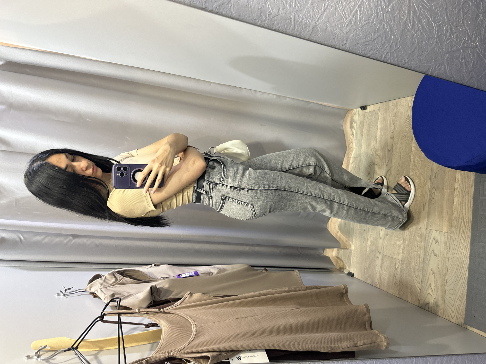 A woman stands in a fitting room, taking a mirror selfie. She is wearing a light beige t-shirt, grey acid-wash jeans, and black and white striped sandals. A small white shoulder bag is slung across her body. In the background, a beige ribbed tank dress hangs on a clothes rack, and grey curtains line the walls. The floor is made of light-colored wood planks. The scene is likely within a retail store, possibly in an unknown city.