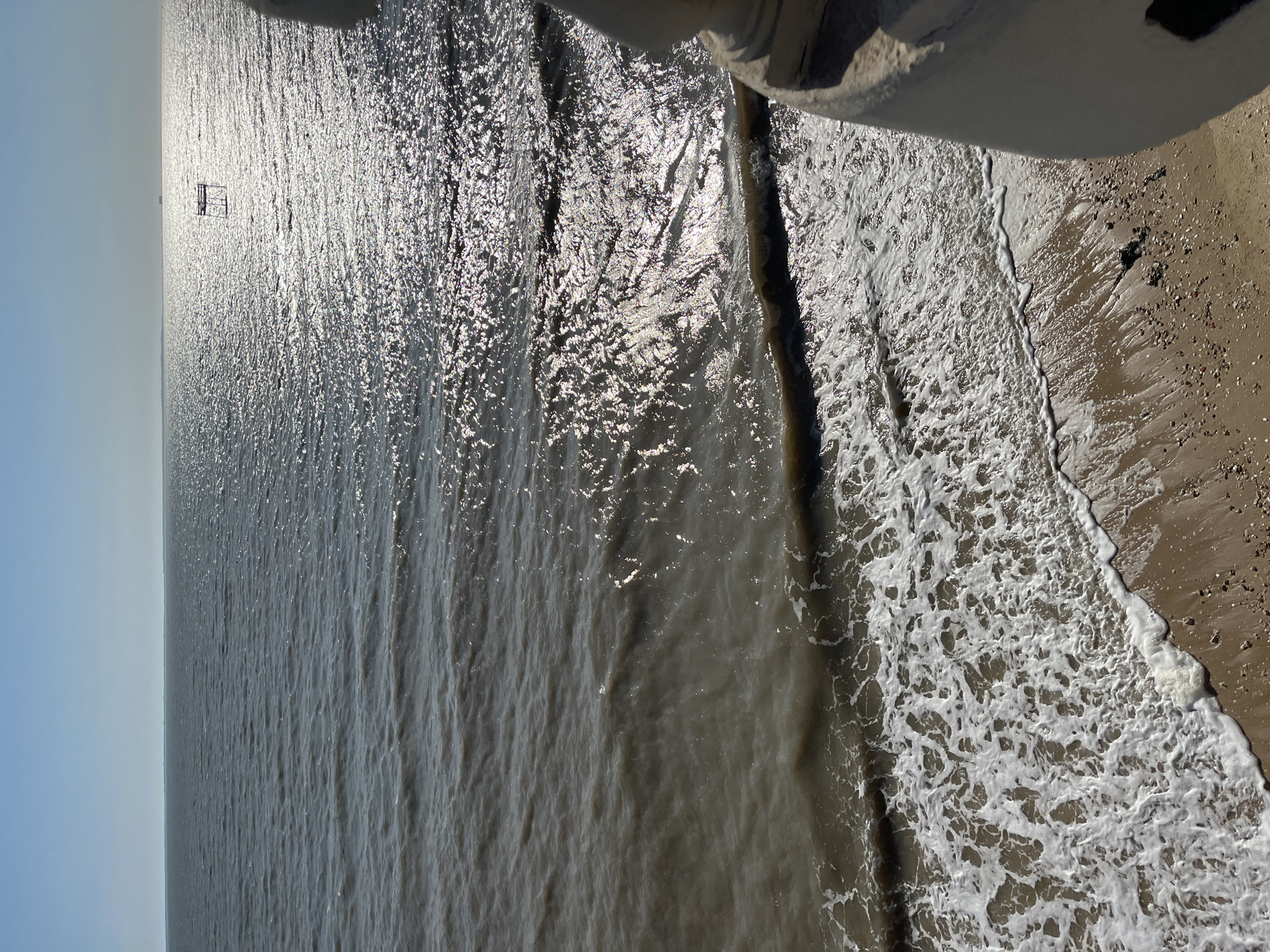 The image shows a view of the sea from a slight elevation, likely a promenade or building overlooking the coast in Fouras, France. The water is a murky brown, with small waves breaking onto a sandy beach. The sun is high in the sky, casting a bright glare on the water's surface. In the distance, a small, man-made structure, possibly a fishing platform or buoy, is visible in the water. To the right of the frame, a portion of a white, possibly stone, railing or wall is visible, suggesting the vantage point is from a man-made structure. The beach itself is composed of sand and small pebbles. No people are visible in the frame. The overall impression is one of a calm, sunny day at the seaside.