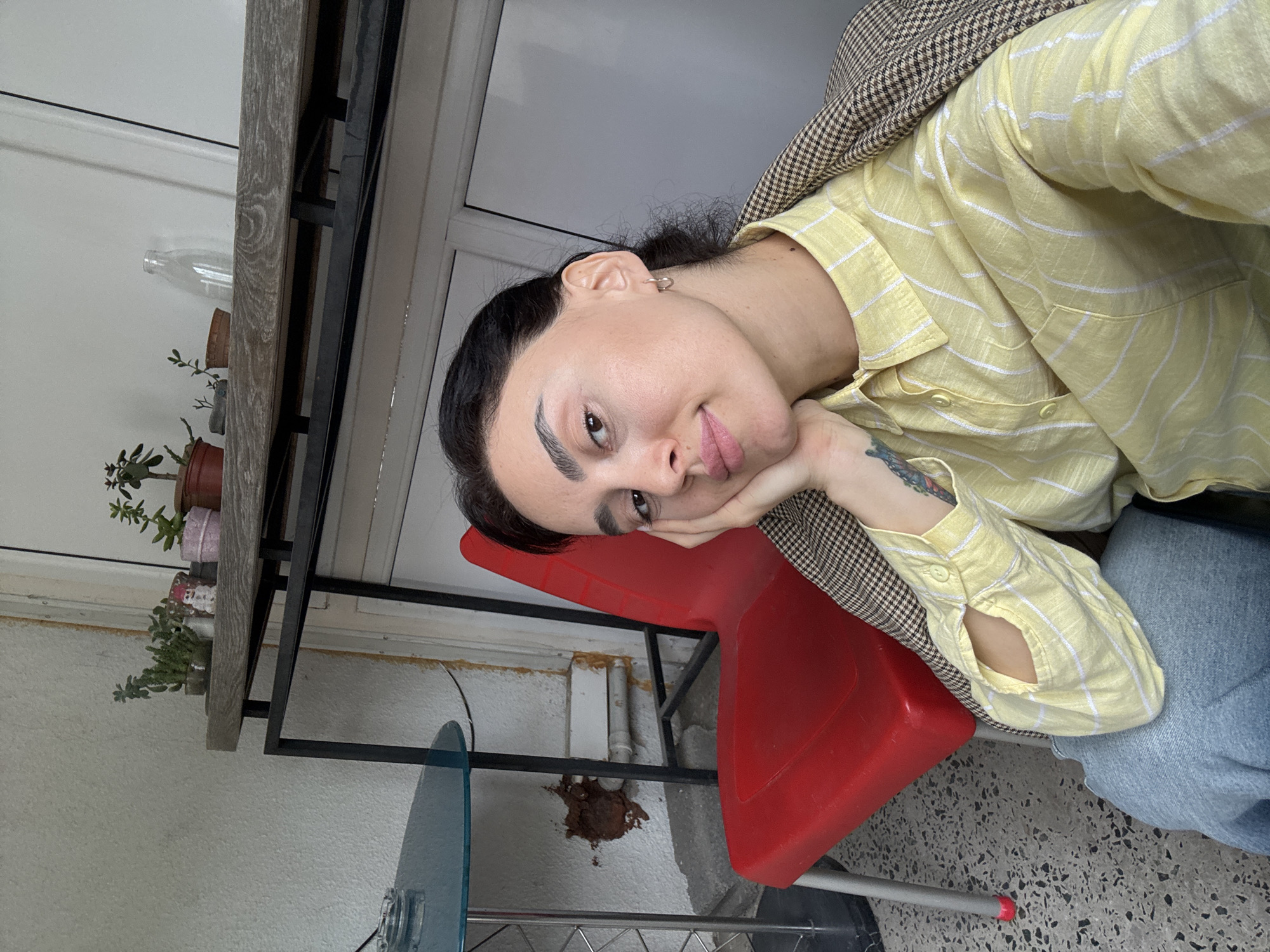 A young adult woman with dark hair pulled back is resting her face on her hand while seated on a red plastic chair indoors. She is wearing a yellow striped shirt and a brown checkered jacket. She is also wearing light blue denim jeans. In the background, there is a table with plants on it, a window, and a glass-topped table. A large patch of brown material is adhered to the wall behind the chair. It is likely located in Tbilisi, Georgia, based on common architectural features, though nothing specifically identifying it to that region can be seen.