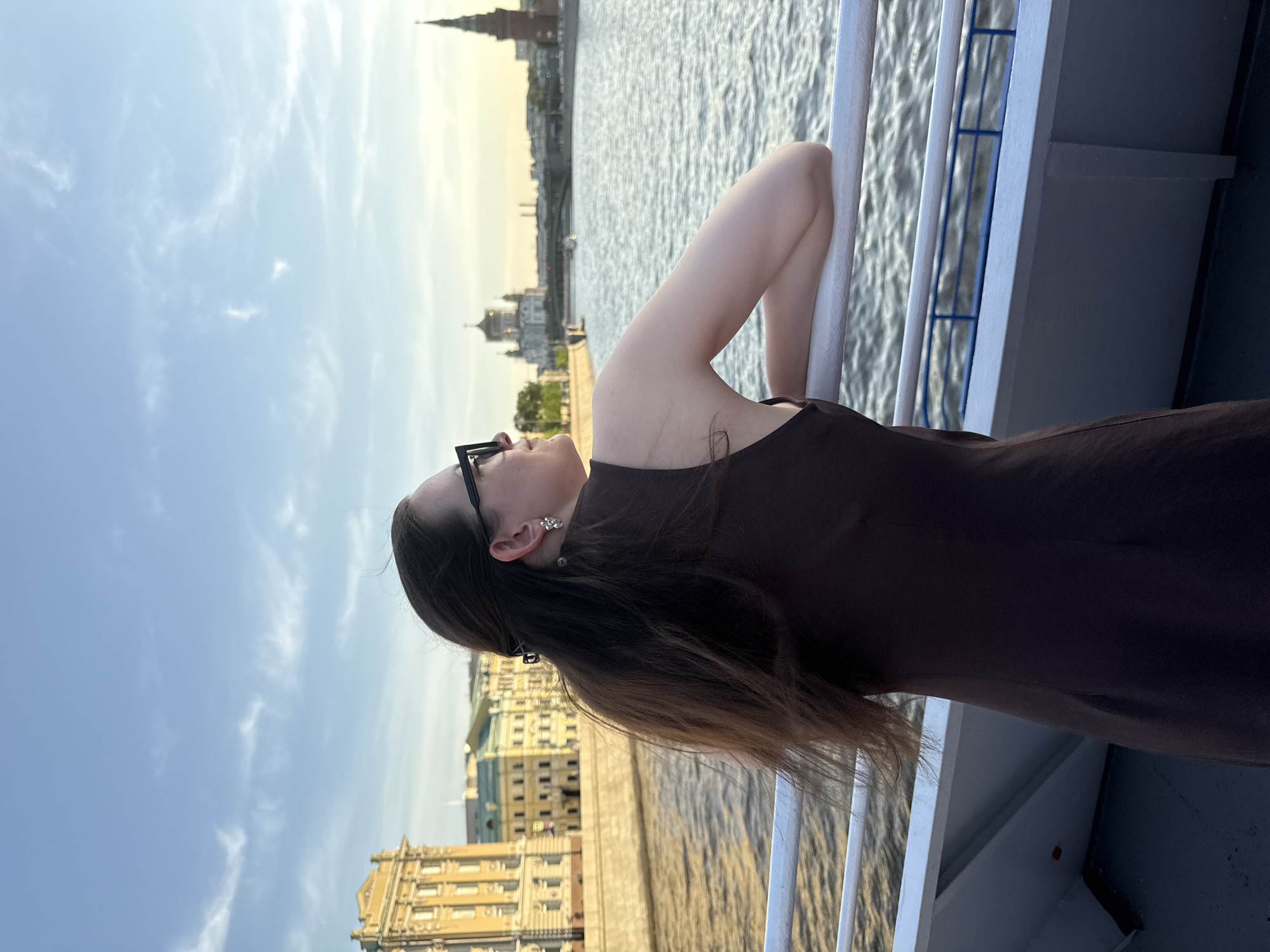 A woman with brown hair and sunglasses stands on a boat in Moscow, Russia. She wears a brown dress and silver earrings. She leans against the white railing of the boat, looking upward. In the background, the Moscow Kremlin and other buildings are visible along the riverbank. The sky is blue with some clouds. The river is visible behind her.
