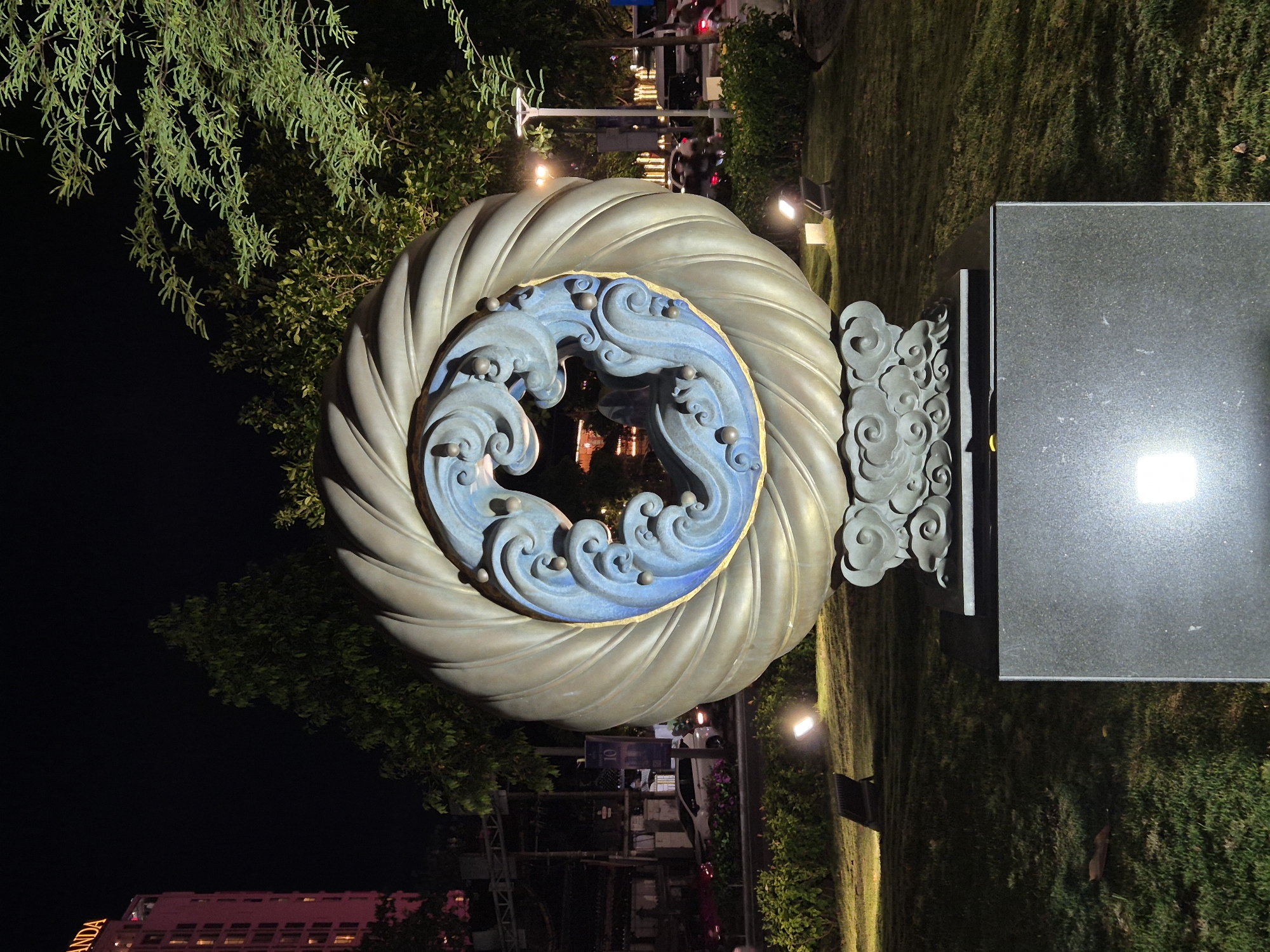 The image shows a large, circular sculpture with a metallic, ribbed outer edge and a blue, wave-like interior. The sculpture sits on a tiered, cloudy base, which in turn rests on a dark granite pedestal. The scene is illuminated at night by various lights, including spotlights on the grass and lights from a nearby building. In the background, a tall building with the illuminated letters "ANDA" is visible on the left. There are trees and streetlights, and cars can be seen on a road in the middle ground. This sculpture is located at the Dragon Bridge in the city of Đà Nẵng, Vietnam.
