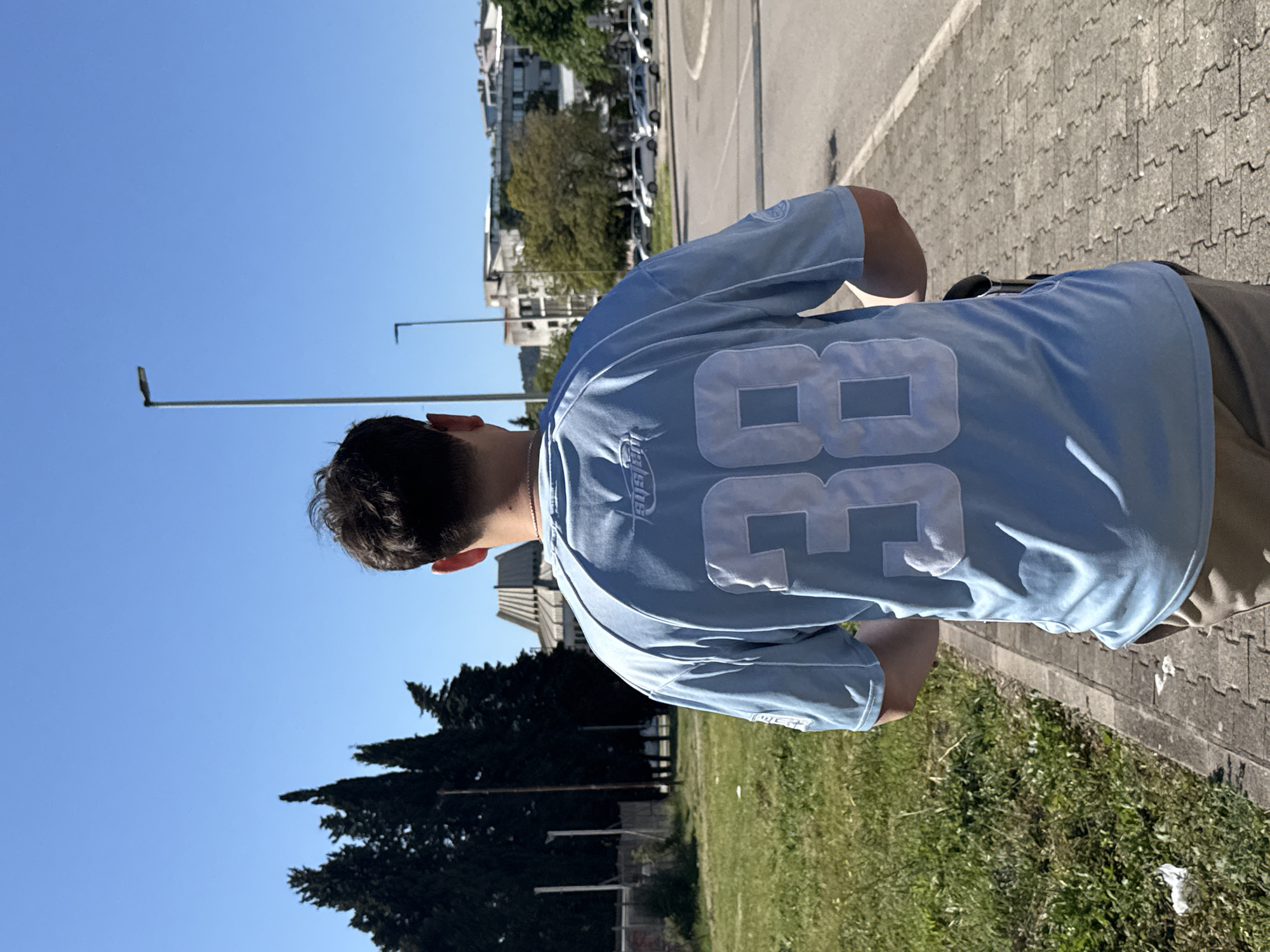 A medium shot captures a person from behind, wearing a light blue jersey with the number "38" in white, positioned in what appears to be a city environment. The subject stands on a paved walkway with green grass and sparse vegetation to the right, and what seems like a parking lot and road on the left, hinting at urban or suburban setting. In the background, buildings and trees suggest an urban landscape. Given the reference to the city of Bar, Montenegro, the surroundings may reflect the region's typical architectural style and vegetation. The image is rotated slightly, which causes the sky to appear on the left, and the ground on the right.
