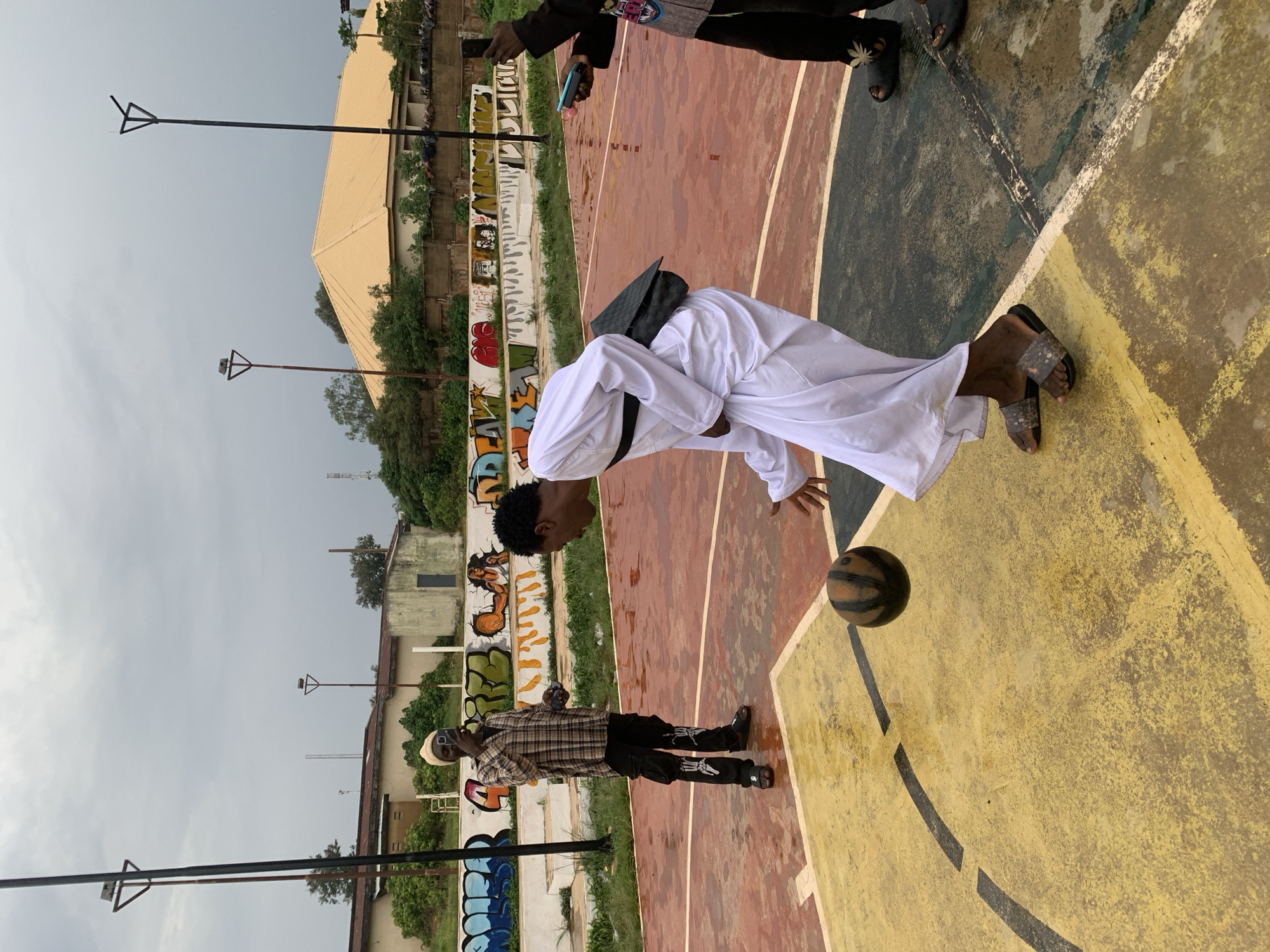 In Jos, Nigeria, on a wet outdoor basketball court, three young men are interacting. The man in the center, wearing a white robe and sandals, is in the process of dribbling a basketball. To his left, a man in a plaid shirt, black pants adorned with skeleton graphics, and a beanie is holding a smartphone, seemingly taking a selfie. To the far right, another man, partially obscured, wears black pants and sandals and holds two phones, one pink and one blue, in his hands. The basketball court's surface is a mix of red and yellow with black markings. In the background, a concrete wall displays colorful graffiti, and beyond that, buildings and trees are visible under a cloudy sky. The scene suggests a casual gathering and playful activity.