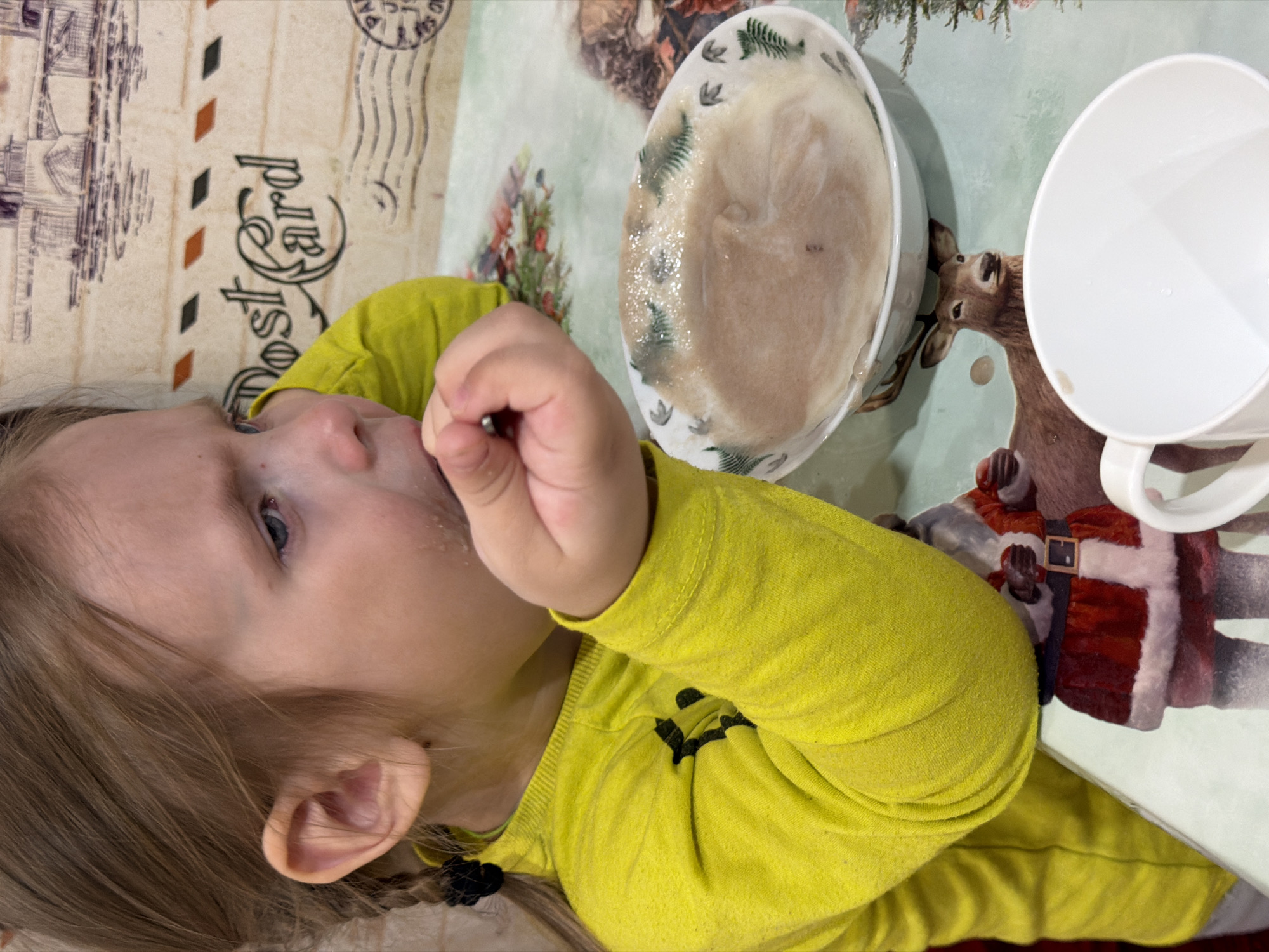 A young child is seated at a table with a bowl of porridge and a white cup. The child is wearing a yellow long-sleeved shirt and has blond hair. The table covering features a postcard design on one side and a winter scene with a deer and Santa Claus on the other. The scene appears to be indoors. This scene is taking place in the city of Chaikovsky, Russia.