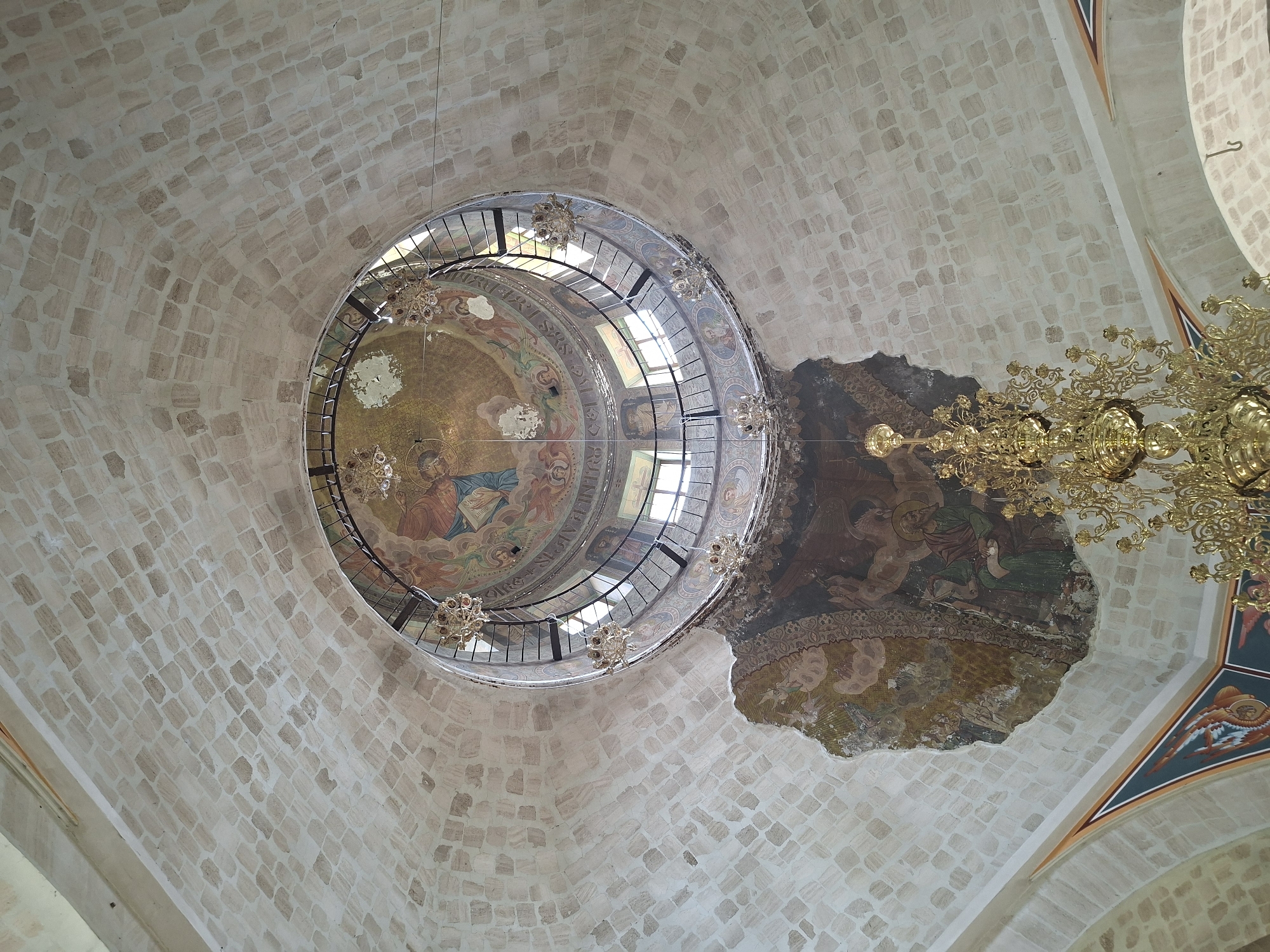 The image shows the interior of a domed structure, likely a church, in Condrița, Moldova. The dome is constructed of light-colored, rectangular stones arranged in a pattern. A circular opening at the apex of the dome is visible, with a metal railing and windows. Below the opening, a circular fresco depicting religious figures in colorful robes is partially visible. Several ornate, gold chandeliers hang from the ceiling. A large section of plaster on the dome has fallen away, revealing a fresco underneath with figures in earthy tones. The frescoed section that remains intact depicts a seated figure in green robes, with a scroll in hand, in front of a decorative border. The overall impression is one of historical architecture with significant artistic and decorative elements, some of which show signs of deterioration.