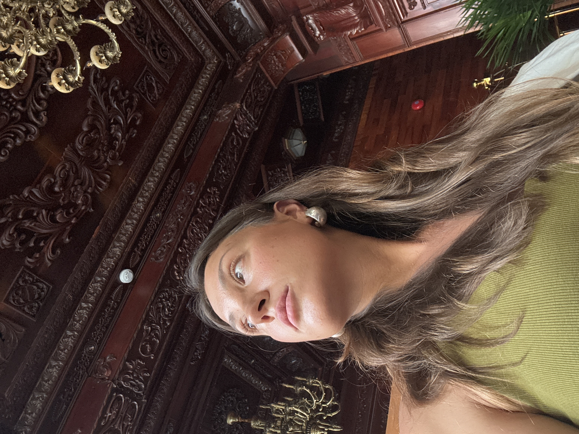 The image shows a woman with brown hair wearing a green shirt indoors. The background features ornate dark wood paneling, decorative molding, and chandeliers. The location is likely a formal room or interior in Đà Nẵng, Vietnam, given the architecture and style of the wood decorations.