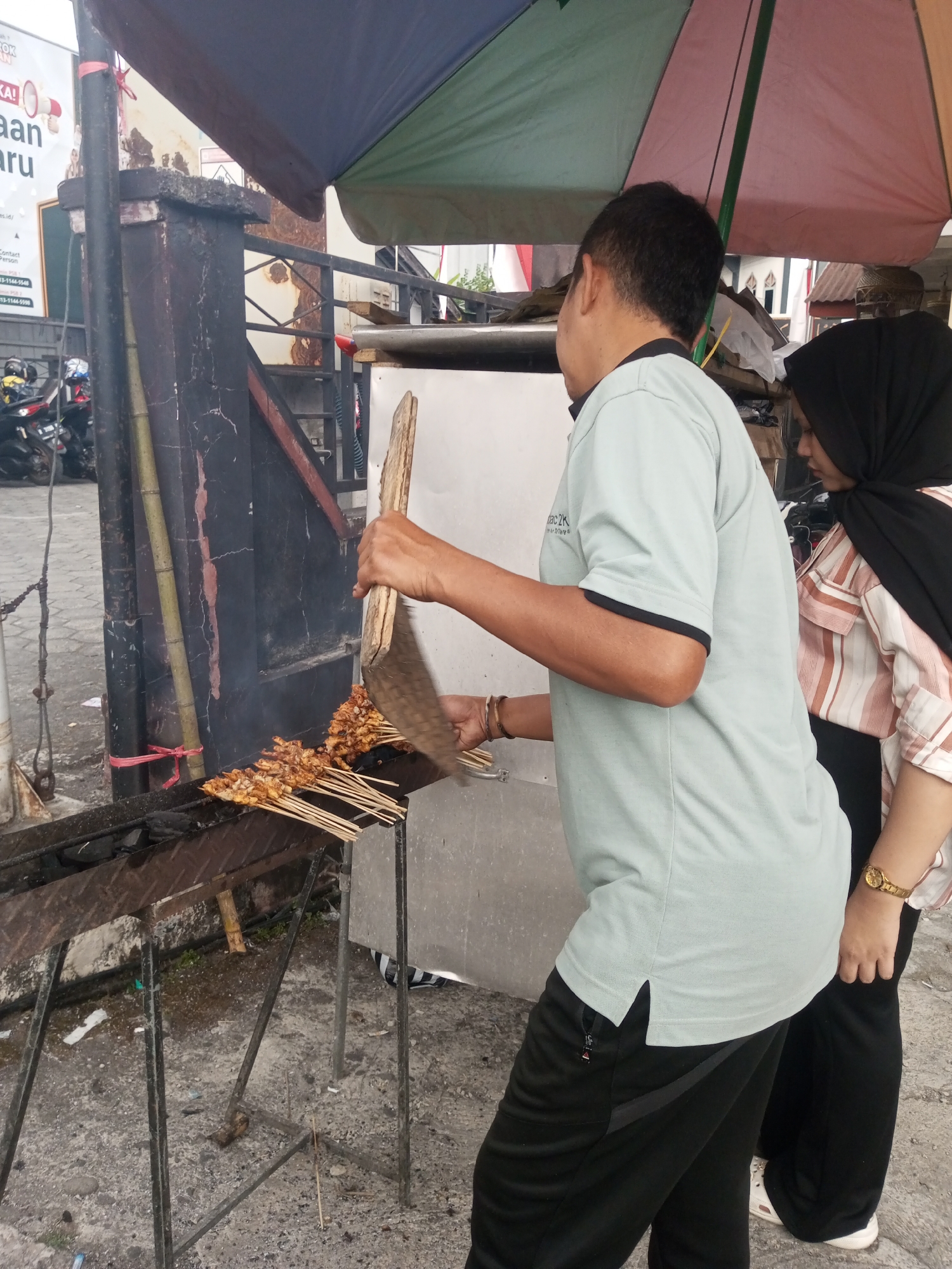 The image depicts an outdoor food stall in Pandansari, Indonesia, where satay is being grilled. A man, wearing a light blue t-shirt and black pants, is actively grilling the satay using a wooden tool to flip the skewers over a charcoal grill. To his right, a woman wearing a black hijab and a striped shirt stands nearby, observing the grilling process. The stall is set up with a metal grill, bamboo poles, and a colorful umbrella providing shade. The ground is paved with concrete and appears somewhat worn. In the background, motorcycles are visible, suggesting the stall is located in a public area, possibly a street or market. The overall scene captures a moment of food preparation and commerce in a bustling Indonesian environment.