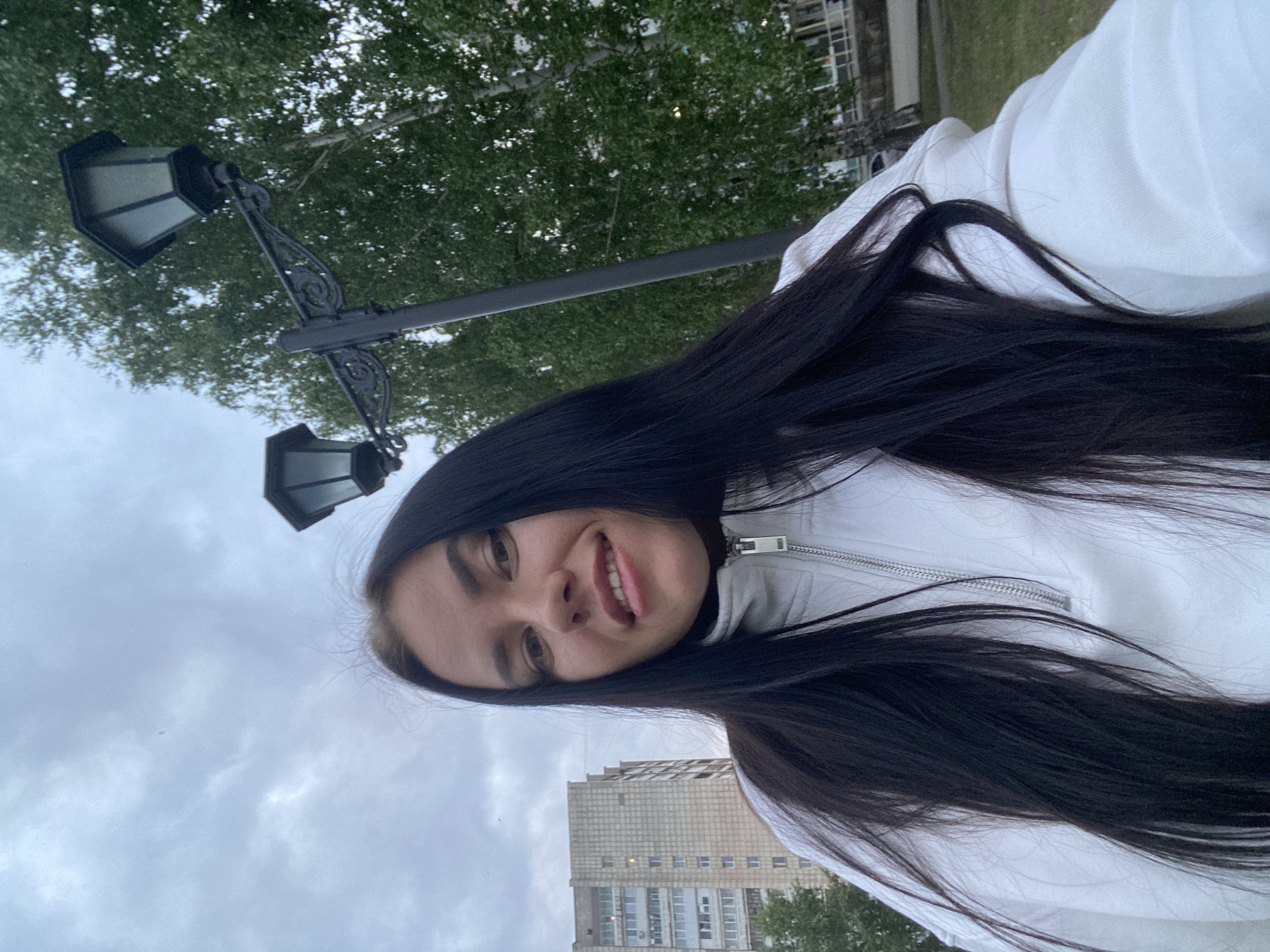 A young woman with long, dark hair is taking a selfie outdoors. She is wearing a white zipped sweatshirt. Behind her, a tall, light-colored apartment building is visible, as well as a street lamp with two lights and a backdrop of green trees and an overcast sky. The image appears to be taken in the city of Chaikovsky, Russia, likely in a residential area.