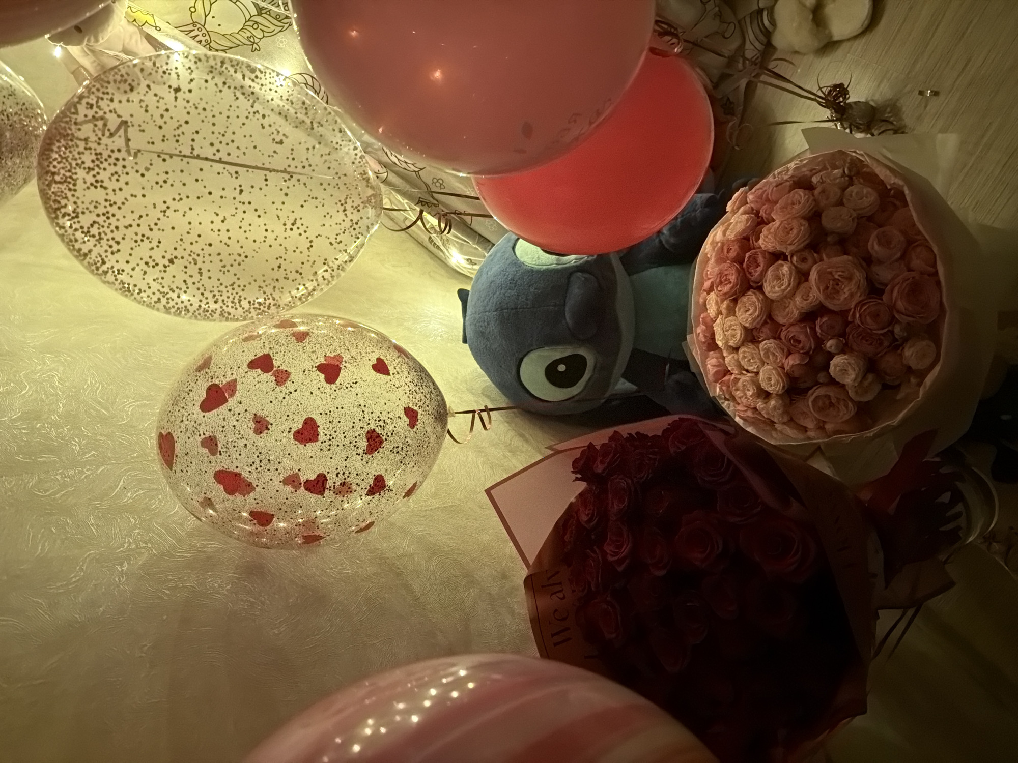 The image shows a close-up, indoor scene with several objects scattered on a surface with a textured, possibly fabric-like, covering. There are three balloons: one with glitter, one with hearts, and two opaque, pink/red balloons. A blue stuffed animal, resembling the character "Stitch," is also present. Two bouquets of flowers, one of red roses and another of smaller, pink roses, are visible. The lighting is warm and somewhat dim. The location in the city of Chaikovsky, Russia is not visible. No people are depicted in the image. The focus is on the arrangement of objects, suggesting a celebratory or gift-giving context.