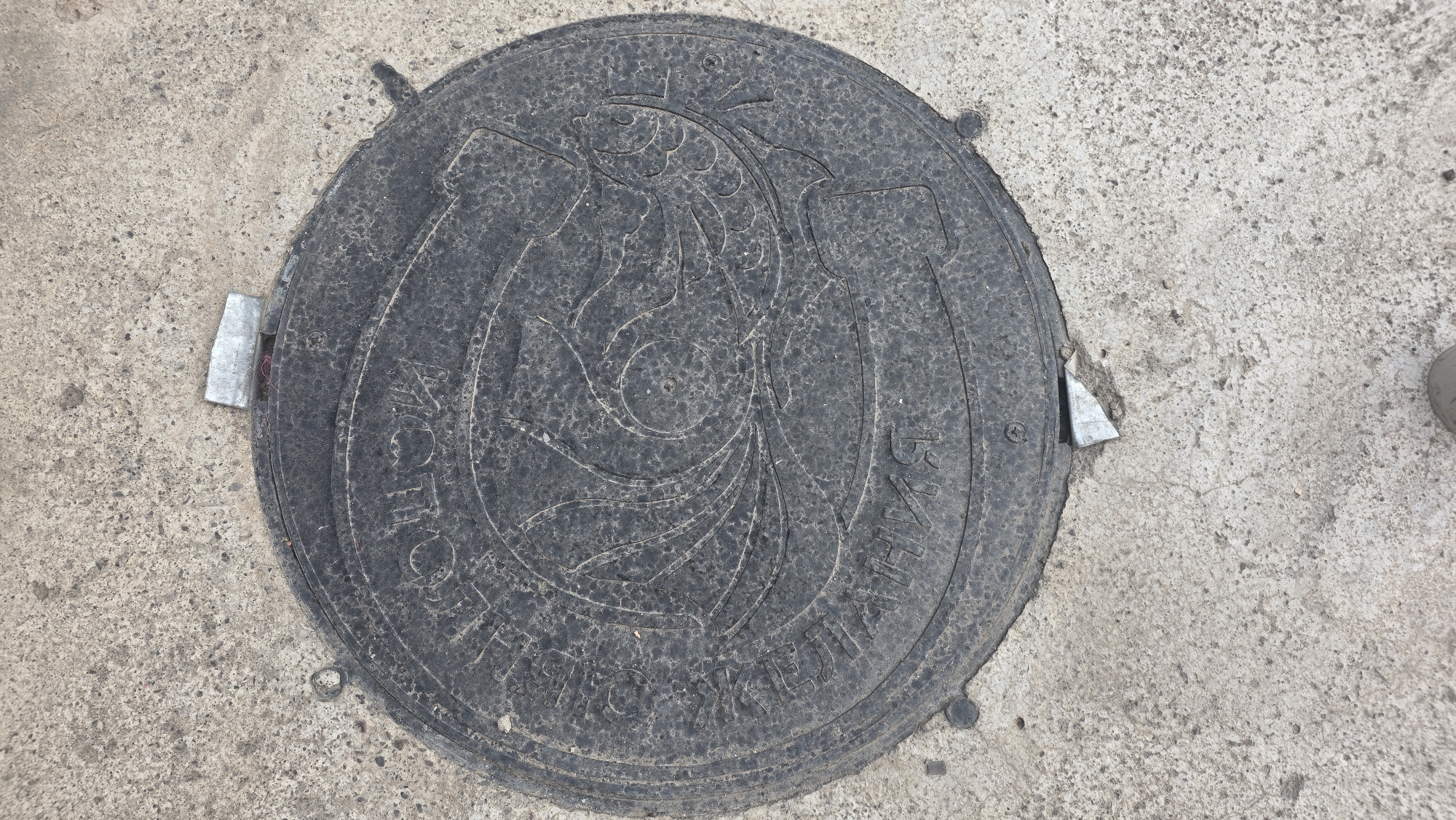 An overhead, medium shot captures a circular, dark metal manhole cover set into a light gray, textured concrete surface. The manhole cover is centrally positioned, slightly angled. Its surface features a raised circular design that appears to be an artistic depiction of a fish swimming amongst wavy lines or plant elements. There is text inscribed in a circular pattern around the outer edge of the manhole cover, though it is largely obscured by dirt and wear. Small, metallic wedges are inserted at two points along the edge of the manhole cover, possibly to secure it. The concrete ground is rough and shows some minor cracks and debris. A portion of a person's shoe is visible in the bottom right corner, indicating a human presence. The scene is likely outdoors in an urban or semi-urban environment, given the presence of a manhole cover and concrete. The text on the manhole cover, if legible, could provide further clues to its origin or purpose within the city of Chuy, Kyrgyzstan.