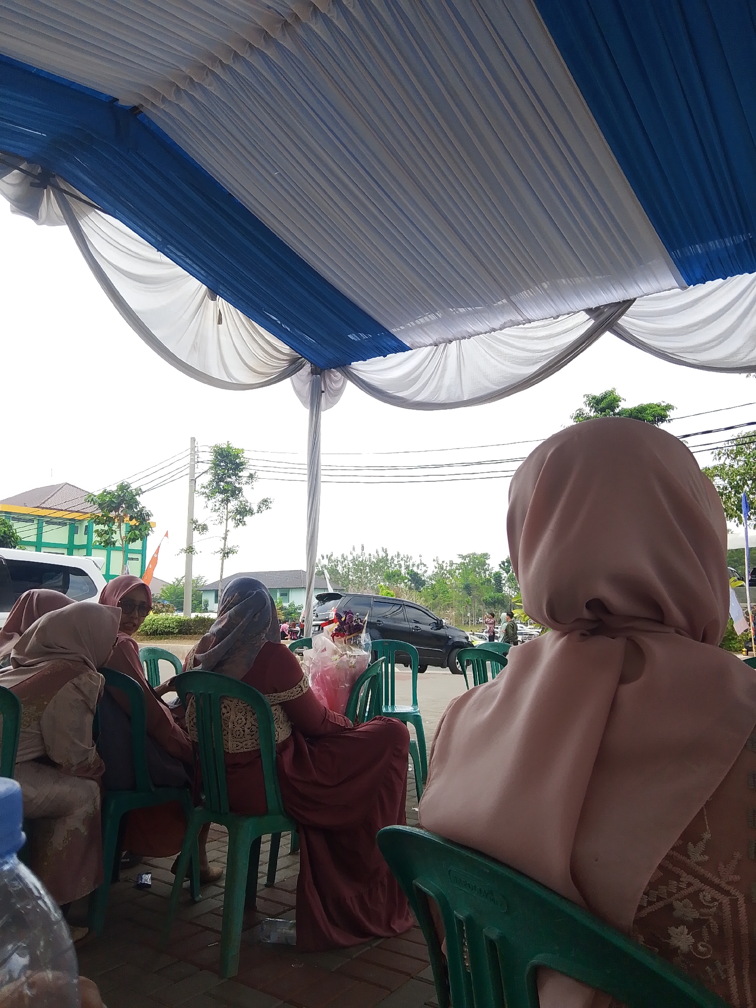 The image depicts an outdoor gathering under a white and blue striped tent. Several women, all wearing hijabs, are seated on green plastic chairs. The woman in the foreground on the right is facing away from the camera, her head and shoulders visible, wearing a light brown hijab and a patterned top. To her left, a woman in a dark red dress and a grey hijab is also seated. Further to the left, two more women in hijabs are partially visible, one in a light brown hijab and the other in a peach-colored hijab. In the background, there are cars, trees, and buildings, including a green and white building with a red flag. The ground appears to be paved with bricks. The overall impression is of a casual outdoor event, possibly a celebration or a community gathering. The image does not contain enough specific visual cues to identify a precise location within Tasikmalaya, Indonesia.