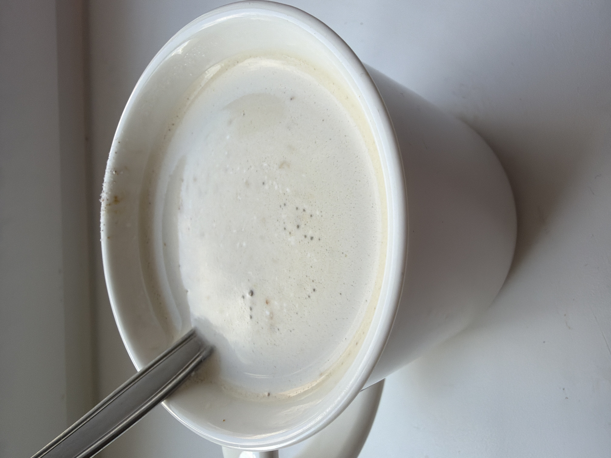 A close-up, eye-level shot shows a white mug filled with a foamy, light-brown liquid, presumably coffee or latte, with a silver spoon partially submerged in it. The mug is placed on a white surface, possibly a windowsill, with a soft, grey background suggesting an indoor setting. The lighting appears natural and diffused, casting subtle shadows. There are no people present in the image. The scene is devoid of any specific landmarks or identifiers that would place it within the city of Chaikovsky, Russia.