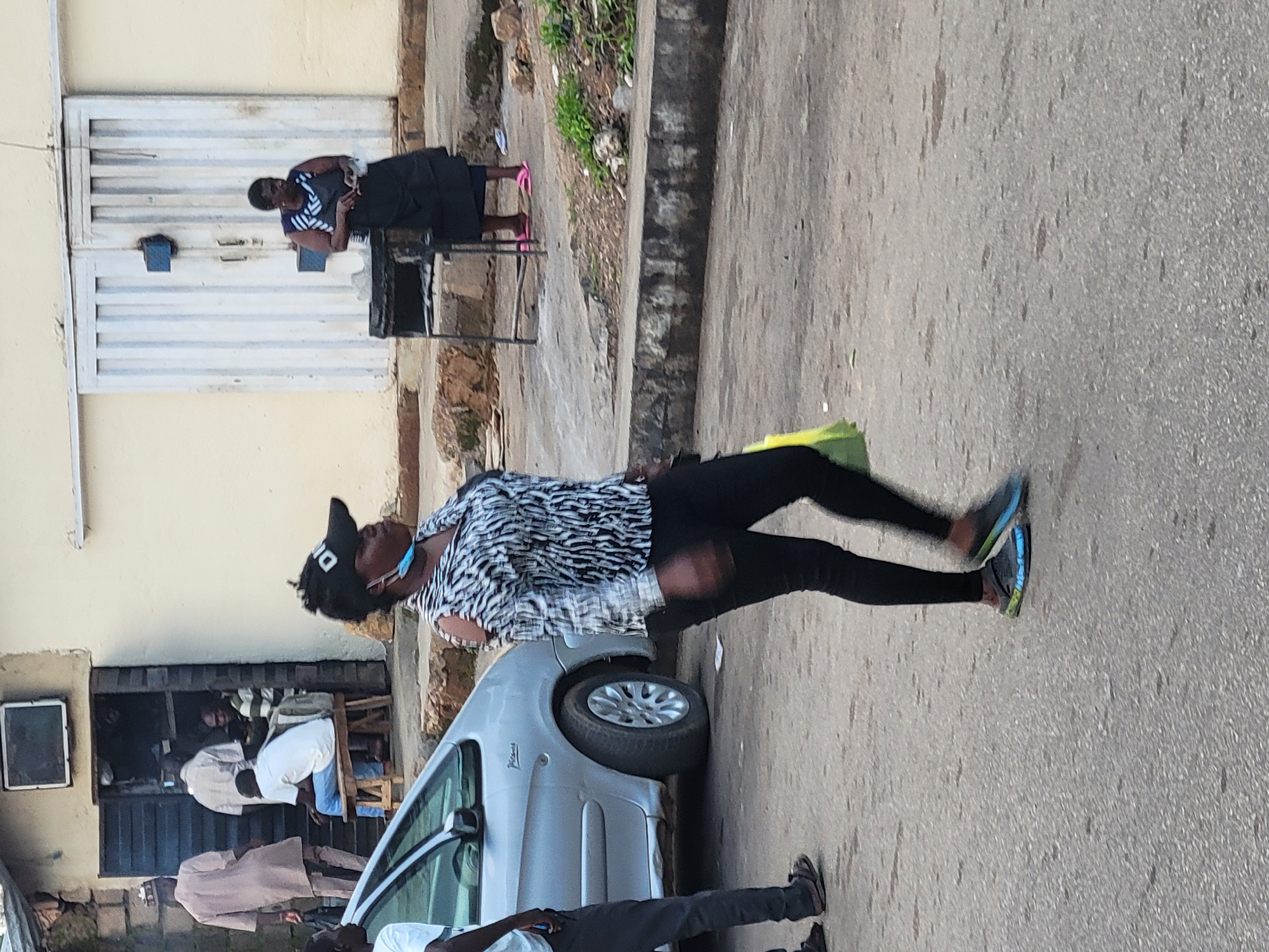 The image depicts a street scene in Barakin Lerawi, Nigeria. In the foreground, a woman wearing a black and white patterned shirt, black pants, and a black cap walks from right to left, carrying a bright yellow bag. A light silver car is parked on the left side of the frame. Behind the woman, several people are visible near a building with an open doorway. One man in a light brown robe stands near the entrance, while another man in a white shirt and blue jeans sits on a wooden stool inside. Further back, a woman in a dark dress and striped top stands near a metal grill structure, facing a large, ribbed white door. The environment appears to be an urban setting with concrete pavement and some debris on the ground.