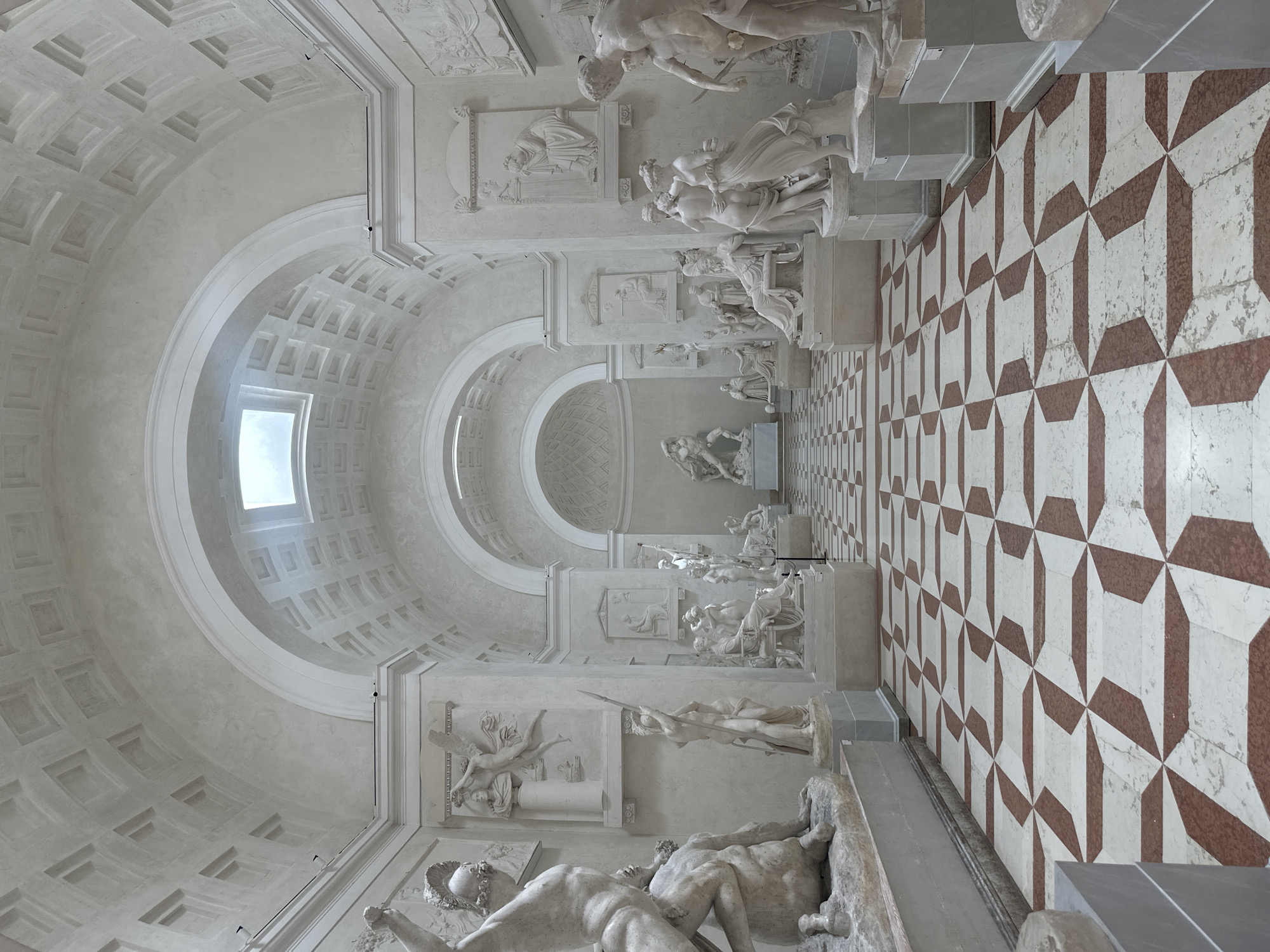 The image depicts a grand, neoclassical-style museum gallery with high, arched ceilings and a skylight. The walls are adorned with bas-relief sculptures and niches containing statues. The floor is paved with a striking geometric pattern of white and reddish-brown marble tiles. Several white marble sculptures of human figures, rendered in classical poses, are displayed throughout the gallery. The overall impression is one of stately elegance and artistic preservation. The location is identified as the Museo Archeologico Nazionale di Cannezza, Cannezza, Italy. There are no people visible in the image.