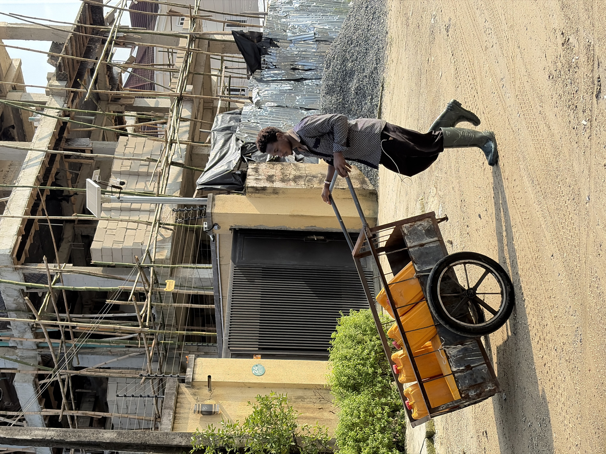 A young man wearing a black and white patterned shirt, dark pants, and green rubber boots is pushing a cart laden with yellow jerrycans. The cart has a metal frame and a single large wheel at the back. The scene appears to be outdoors on a dirt or gravel surface, with tire tracks visible.
In the background, a building under construction is visible, characterized by extensive bamboo scaffolding and exposed brickwork. There are also piles of building materials, including what looks like gravel and sheets of corrugated metal or reflective material covered with black tarps. A solar panel and a security camera are mounted on the side of a building to the left. The sun is casting distinct shadows, suggesting it is daytime. The man's posture and grip on the cart indicate he is actively engaged in moving the load. The overall setting appears to be a construction site or an area where building materials are being stored and transported, possibly in a developing area. No visible text is decipherable in the image.