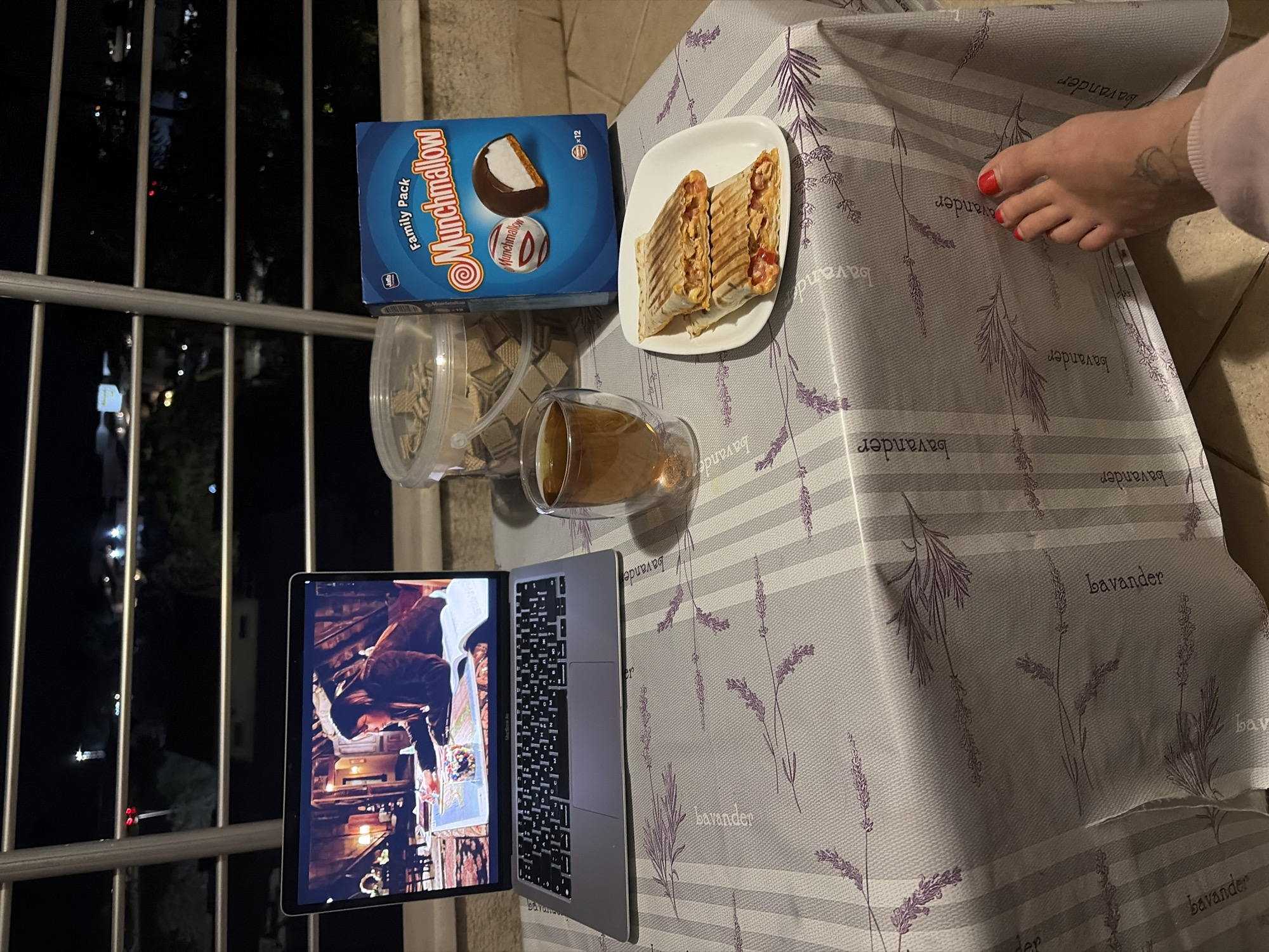 The scene depicts a nighttime still life arranged on a table, seemingly on a balcony. The table is draped with a lavender-printed tablecloth that has the word "bavander" printed on it. A laptop displaying a scene is positioned to the lower left. To the upper left, there's a blue box of "Munchmallow" cookies and a clear container filled with what appear to be wafer cookies. A plate with two halved, grilled panini-style sandwiches rests near the box. Adjacent to the laptop is a glass of liquid, likely a beverage. Finally, in the upper right corner, a human foot with red nail polish is partly visible, suggesting someone is seated nearby. It is not possible to determine the location in the city of Budva, Montenegro from this image.