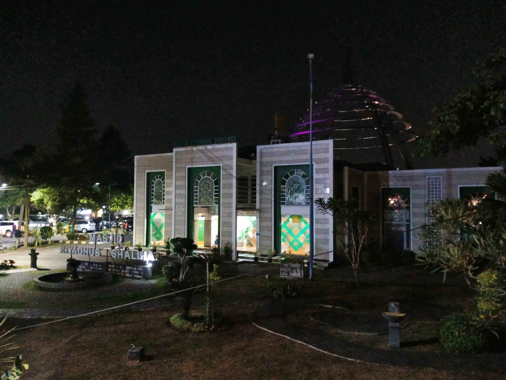 This is a nighttime, eye-level, medium shot of the Masjid Riyadhus Shalihin in Bandung City, Indonesia. The mosque features a modern architectural style with a large, tiered, dome-like structure in the background illuminated by purple and white lights. The main building of the mosque is light-colored with prominent green arched windows and doorways. Inside, warm lighting is visible, illuminating the interior. In front of the mosque, there is a sign with the mosque's name in large, illuminated letters. The foreground consists of a manicured lawn with scattered trees and shrubs, and a small fountain. A paved pathway winds through the lawn. In the background, beyond the mosque, a parking lot with several vehicles and some trees are visible. The overall scene is well-lit by the mosque's artificial lighting against the dark night sky.