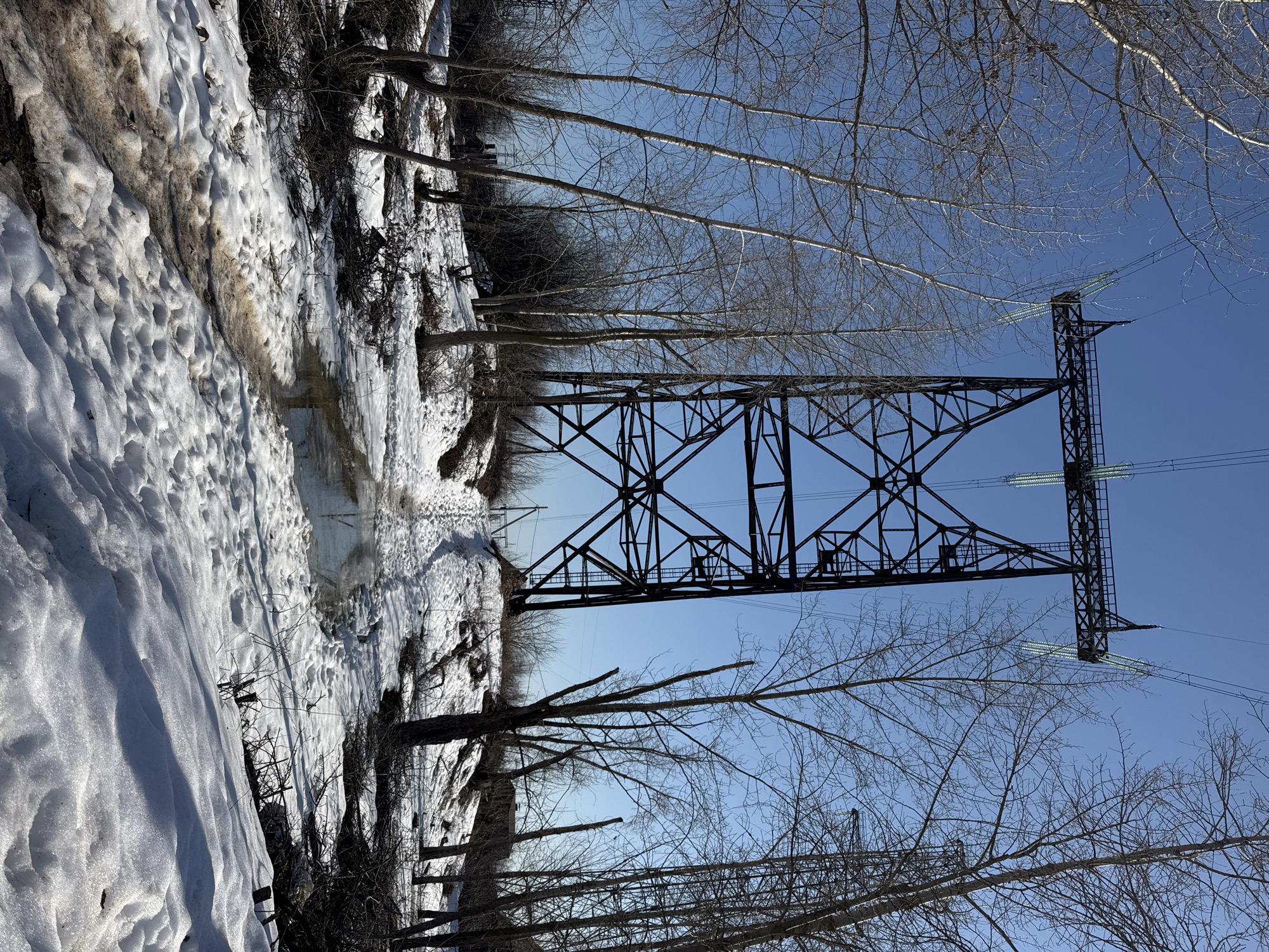The image depicts a winter scene in Chaikovsky, Russia. A metal high-voltage transmission tower stands prominently against a clear blue sky. The ground is covered in snow, with melting patterns and water channels visible. Bare trees line the area around the tower, adding to the winter landscape. The scene appears to be taken from a low angle, emphasizing the height of the tower and the expanse of the sky.