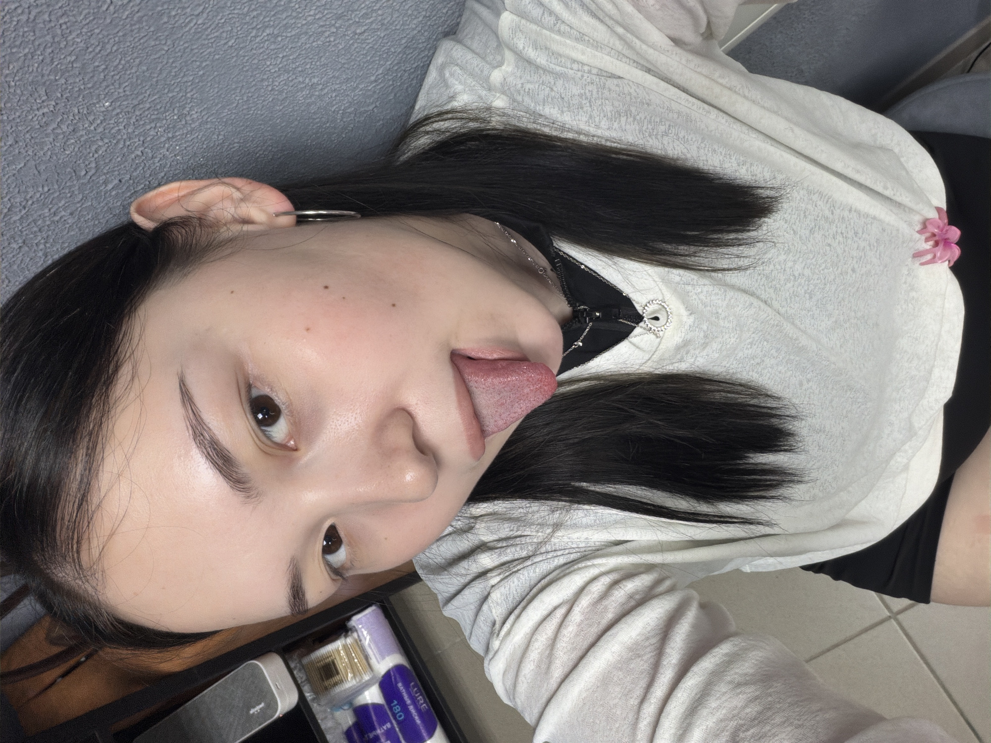 In a close-up, high-angle shot, a young woman with dark hair and light skin sticks out her tongue. She is wearing a white V-neck shirt over a black top and a delicate silver necklace with a teardrop pendant. A silver hoop earring is visible in her left ear. She is positioned in front of a grey textured wall. To her left, a shelf holds various items, including what appear to be cosmetic products and wooden sticks. A pink hair clip is attached to the lower portion of her shirt. The floor appears to be tiled. The context of the city of Chaikovsky, Russia, is not discernible from the image itself.