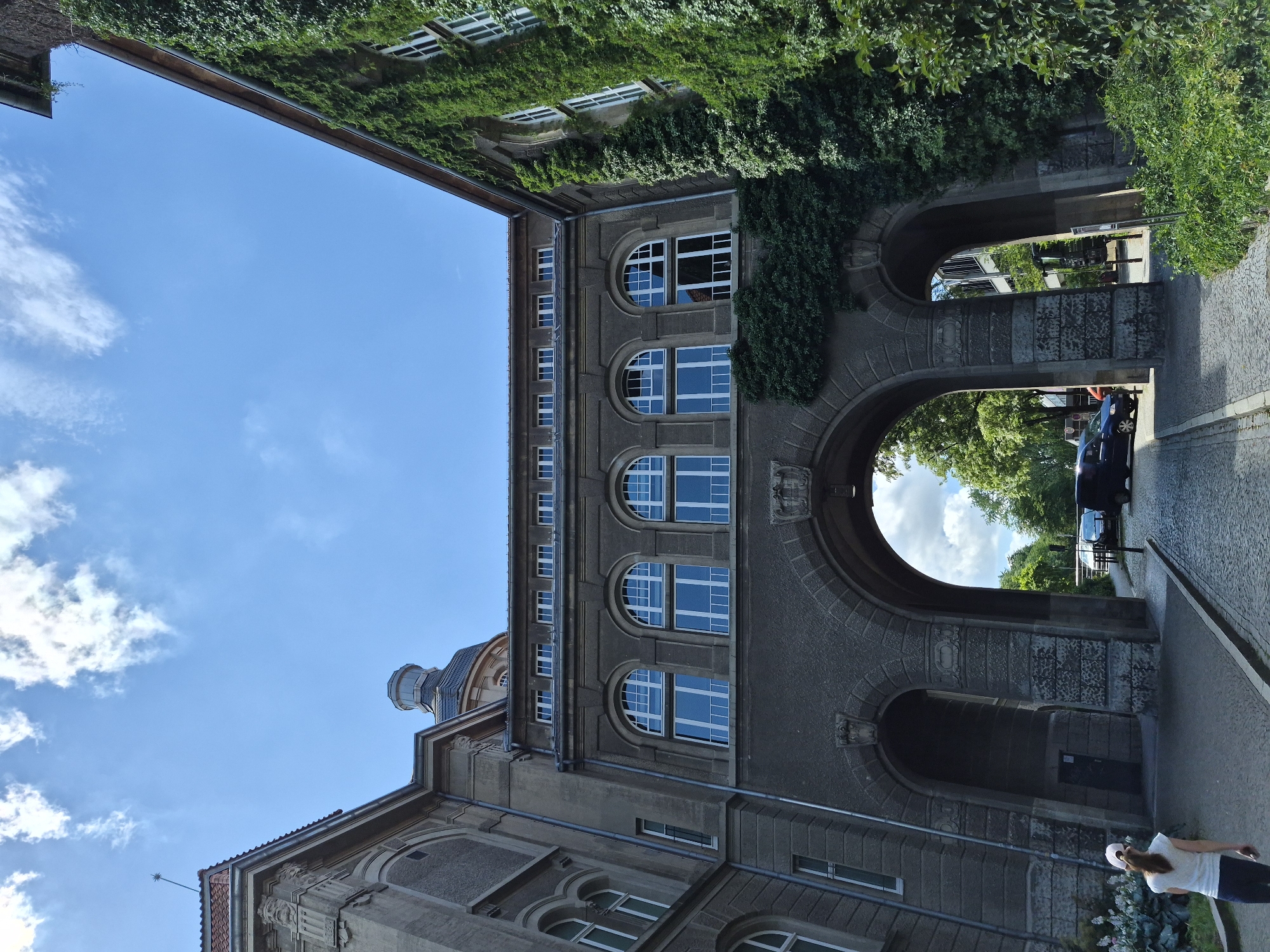 This media file depicts an upward-angled view of an architectural setting in Brunswick, Germany. The primary subjects are the facades of buildings, characterized by their stone construction, arched walkways, and numerous windows. Lush green ivy covers portions of the building exteriors, suggesting an established setting. The scene is illuminated by daylight, with a clear blue sky and some scattered clouds visible, indicating fair weather.
The composition highlights the interplay between the built environment and nature, with the ivy and surrounding trees framing the architecture. Through the arches, glimpses of more trees and a blue vehicle are visible, hinting at a street or courtyard area. A woman in a white t-shirt and dark pants is partially visible in the lower right corner, walking away from the camera, adding a human element to the scene. The architectural style suggests a historical or institutional building. There is no visible text within the image. The perspective and the lush greenery create a sense of depth and a somewhat serene atmosphere.