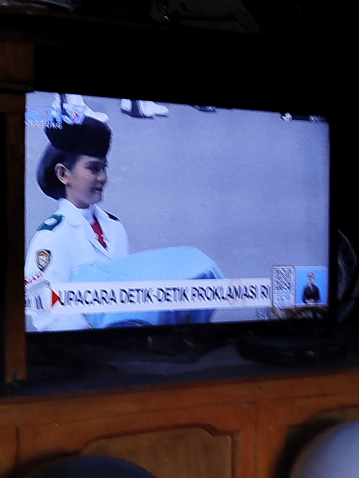 The image displays a television screen showing a broadcast of what appears to be an official ceremony. In the foreground on the screen, a young woman in a white uniform with a black cap is visible. She is wearing a red tie and has a crest on her left shoulder. She is looking to her right. A blue sheet is draped in front of her. Across the bottom of the screen, a banner reads "UPACARA DETIK-DETIK PROKLAMASI RI", which translates to "Ceremony of Proclamation Minutes RI" in Indonesian, suggesting a national event. To the right of the banner, a QR code is displayed, and further to the right, a small inset shows a man in a dark suit. The television screen is displaying a light blue background. The surrounding environment is dark, with wooden furniture visible in the lower part of the image, suggesting the television is in a dimly lit room. The text "RRI" is partially visible in the upper left corner of the screen, indicating the broadcast may be from Radio Republik Indonesia. The scene is consistent with being viewed in Balikpapan, Indonesia, given the Indonesian language text on the screen, which is the national language of Indonesia.