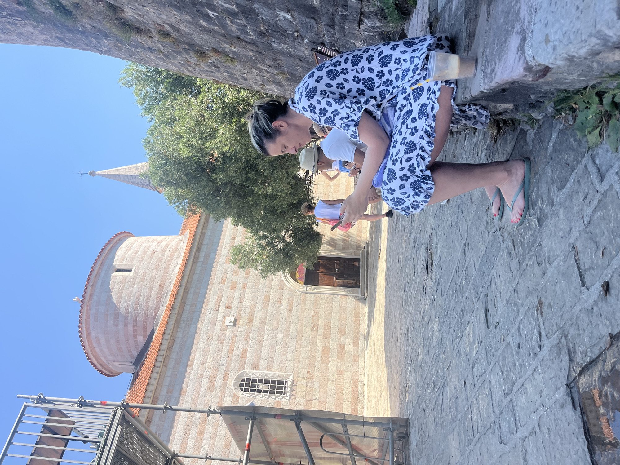 In an outdoor scene on a sunny day, a woman in a blue and white floral dress is squatting on a stone walkway. She has her hair pulled back and is looking down. Beside her, a plastic cup with a straw rests on a stone ledge. Her feet are bare, and she is wearing green flip-flops with pink nail polish. In the background, a stone building with a red-tiled roof is visible, along with a church spire. Trees are also present, and scaffolding is set up in the foreground. Other people are visible in the background, including one wearing a hat. The clear blue sky suggests it is daytime. The scene appears to be in a historic urban area.
