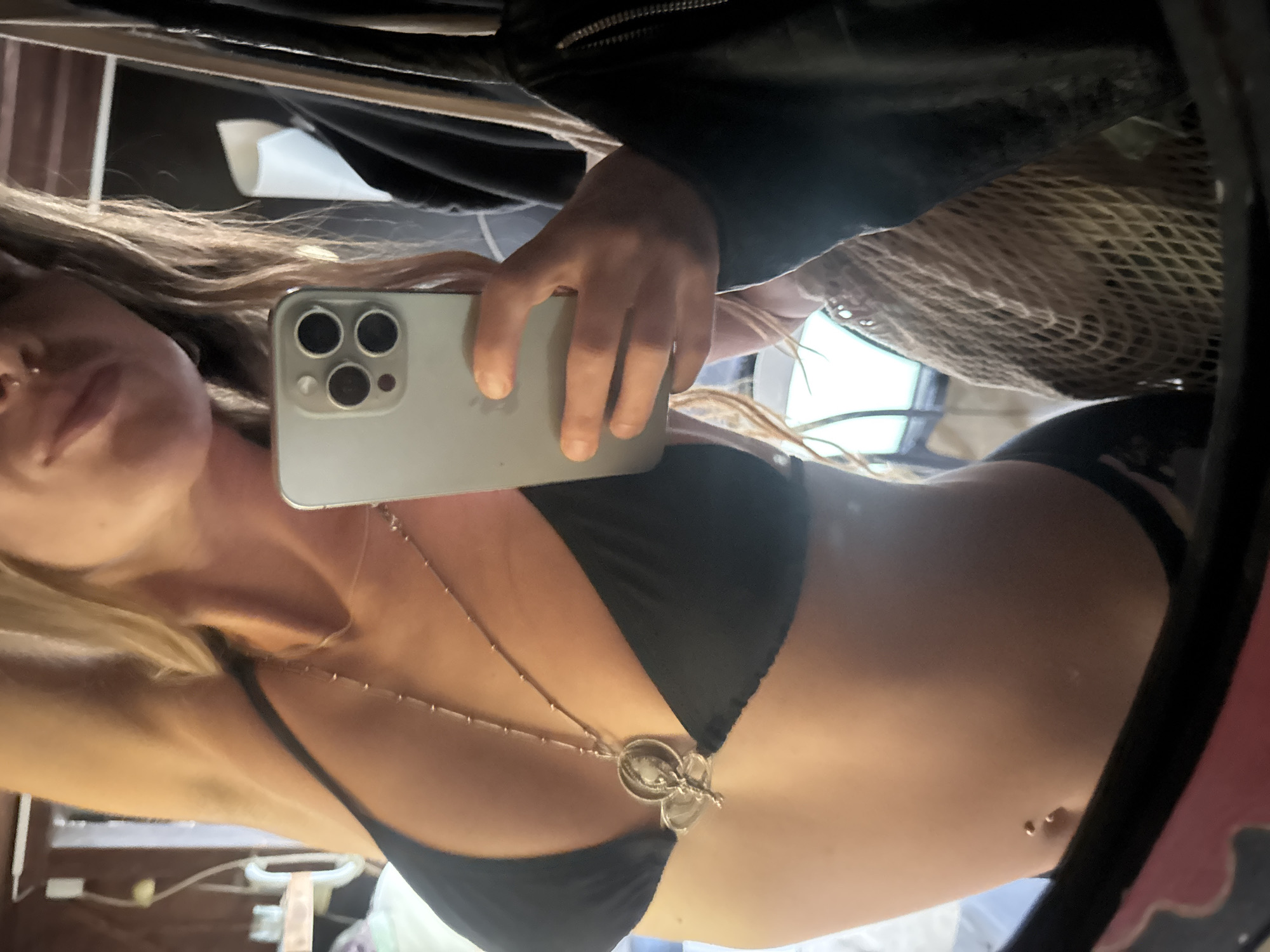 A woman takes a selfie in a mirror. She is wearing a black bikini top and a necklace with a pendant. Her hair is blonde and wavy. She has a belly button piercing. In the background, there are some clothes hanging and a woven bag. The location is an indoor setting, possibly a bedroom or dressing room.