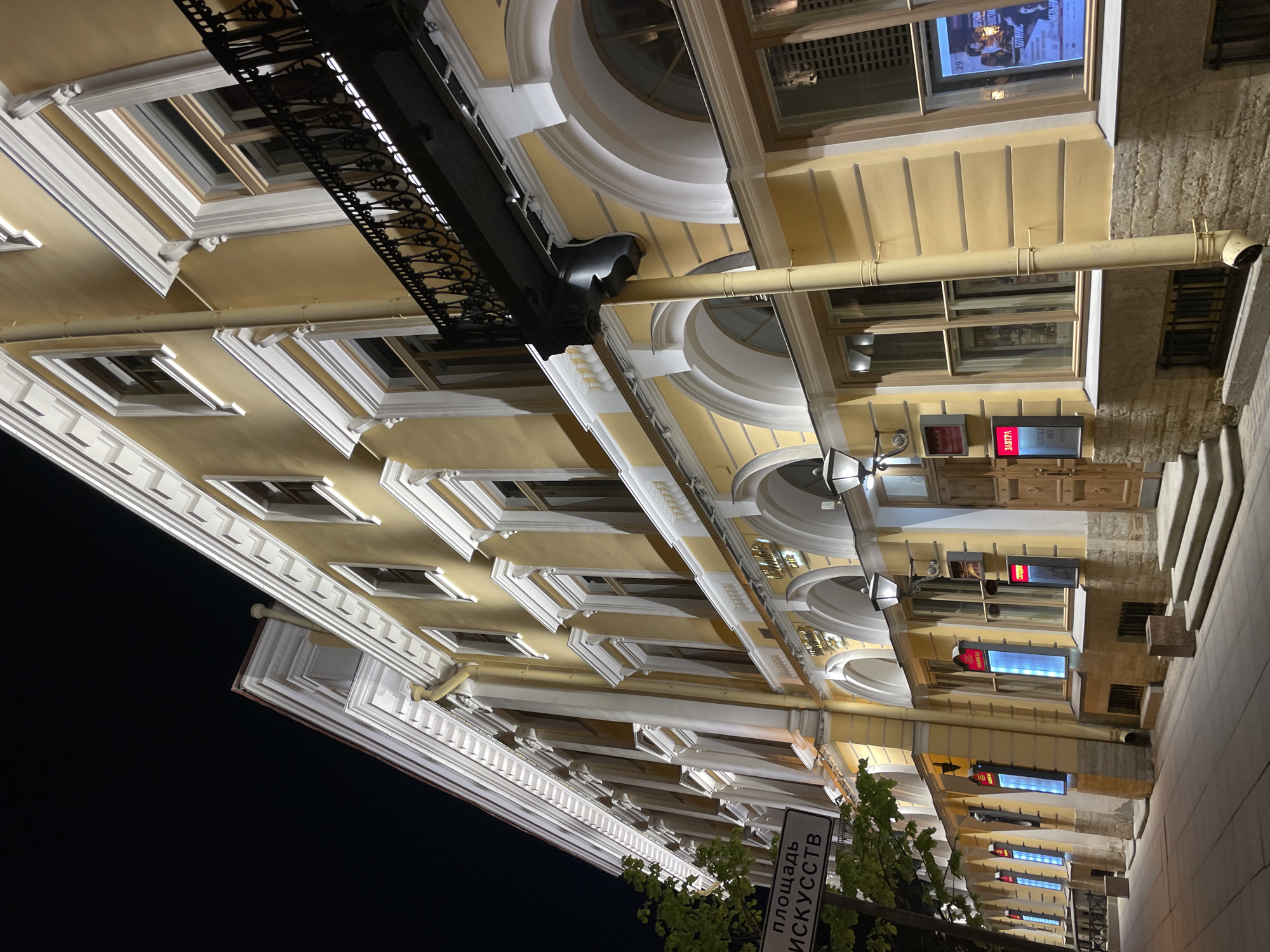 A low-angle, rotated, color photograph depicts a building facade in Saint Petersburg, Russia. The building is primarily yellow with white architectural details, including framed windows and decorative moldings. The facade features both rectangular and arched windows. A black, ornate balcony extends from the building. A drainage pipe runs along the side of the building. Signage with Russian text is visible in the foreground. The sky is dark, suggesting a night scene. The ground and steps are visible on the right. The architectural style suggests a historic or cultural building.