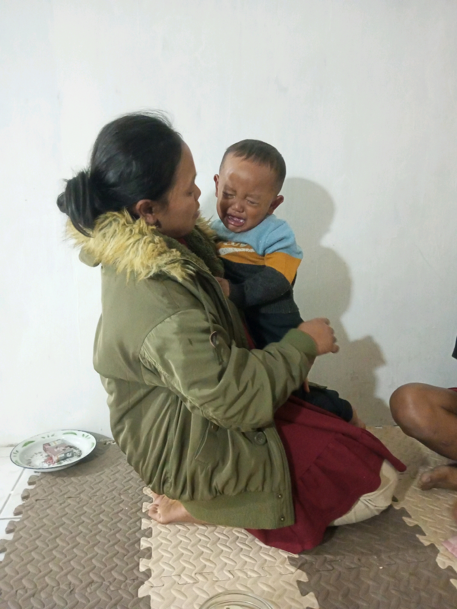 In Marongsari, Indonesia, a woman kneels on a puzzle mat, holding a crying toddler. The woman wears a green jacket with a fur-lined hood and a red dress. The toddler, wearing a blue and yellow shirt and dark pants, is held close to the woman's chest, with its face contorted in distress. Behind them, a white wall casts a shadow. To the left, a small white bowl with items inside sits on the floor. To the right, a person's legs and feet are visible, suggesting another individual is present in the scene. The overall atmosphere is intimate and possibly one of comfort being offered to the upset child.
