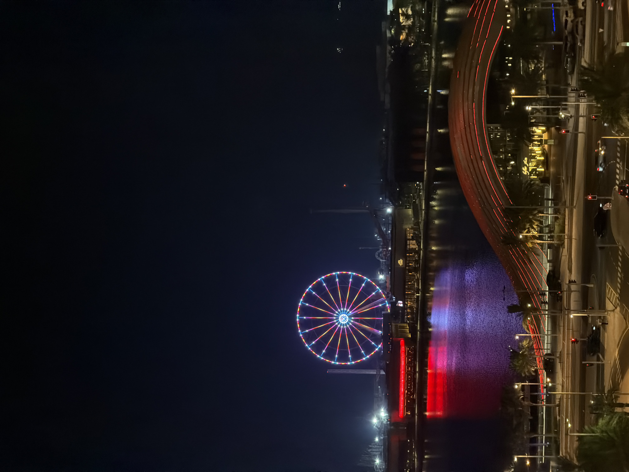 An aerial, long-shot, nighttime photograph captures a cityscape illuminated by various lights. The dominant feature is a large, brightly lit Ferris wheel in the mid-left of the frame, displaying a vibrant array of colors including red, blue, and yellow. In the middle ground, a body of water reflects the colorful lights from the buildings and the Ferris wheel, with a prominent red and purple glow. A modern, arched bridge with red lighting spans across the foreground, crossing over a street. Buildings line the waterfront, with some featuring bright red and yellow lighting. Palm trees are visible along the waterfront and near the street. A street with cars and traffic lights is present in the lower portion of the image, with a pedestrian crossing visible. The sky is dark and clear, characteristic of a nighttime scene. The location is identified as Unknown, Unknown.