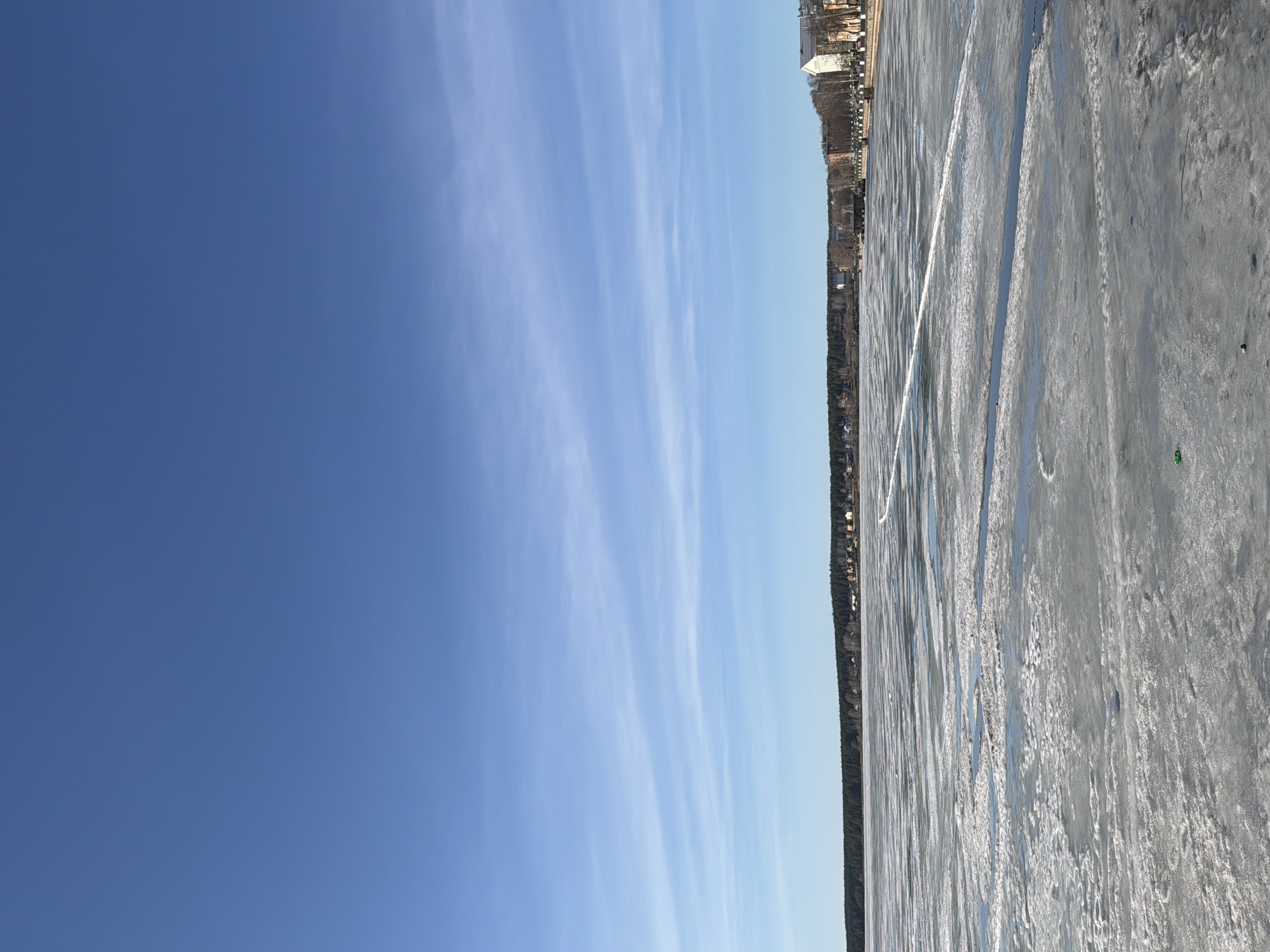 The image shows a view of a frozen body of water, possibly a lake or river, in Votkinsk, Russia. The ice is gray-white, cracked, and has visible markings or tracks on its surface. A treeline runs along the horizon, with buildings visible in the background. The sky is a clear blue with some streaks of clouds. The image is taken during daylight hours with good visibility.
